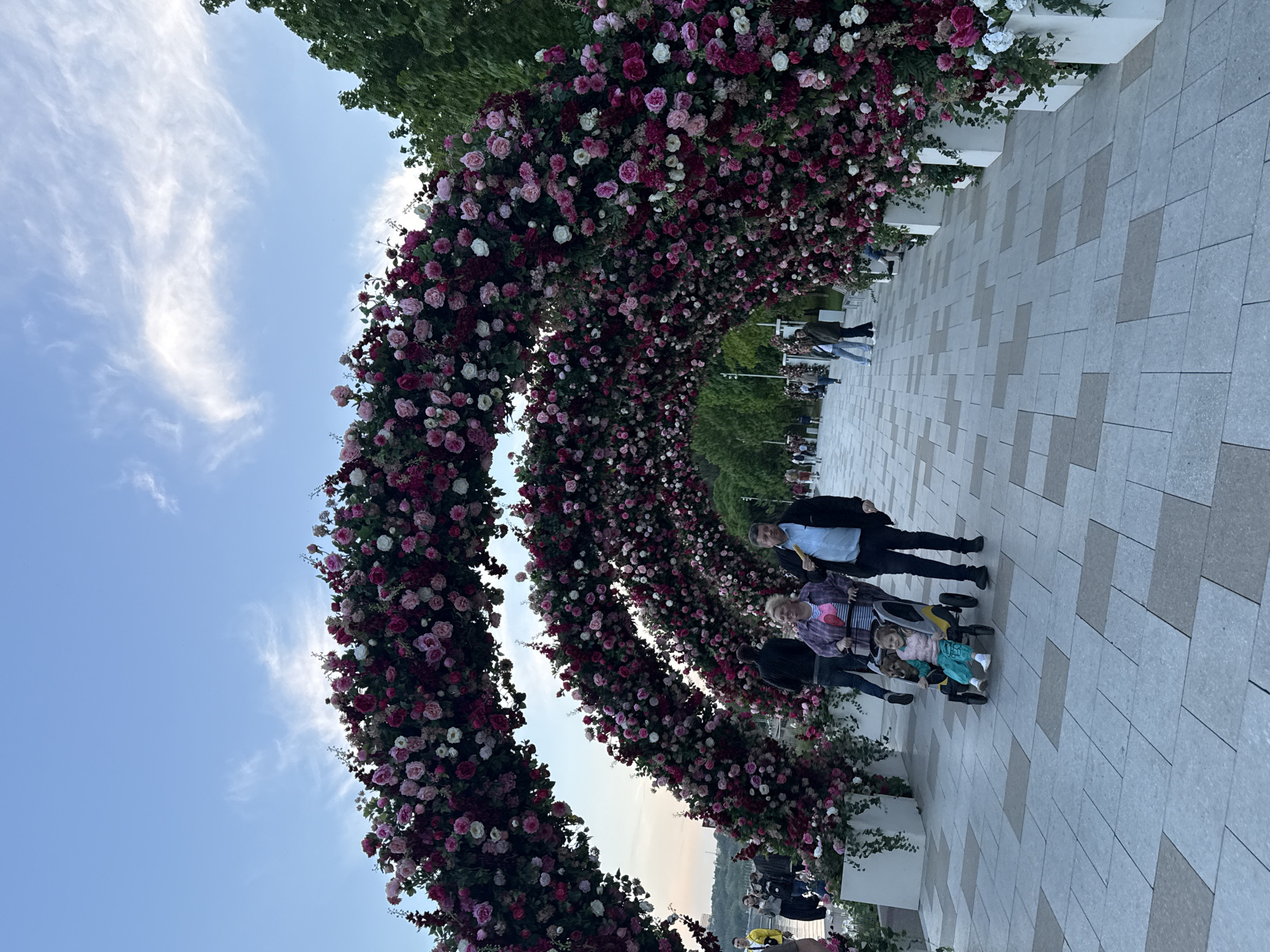 The image shows an outdoor setting featuring a series of arches densely covered with pink, white, and dark red roses. People are walking beneath the arches, including a family with a child in a stroller. The ground is paved with large, light-colored tiles. The sky is visible between the arches, with a blue gradient and some wispy clouds. Trees and greenery can be seen in the background. The scene is recognizable as Zaryadye Park in Moscow, Russia, based on the distinctive rose arches and architectural style of the park.