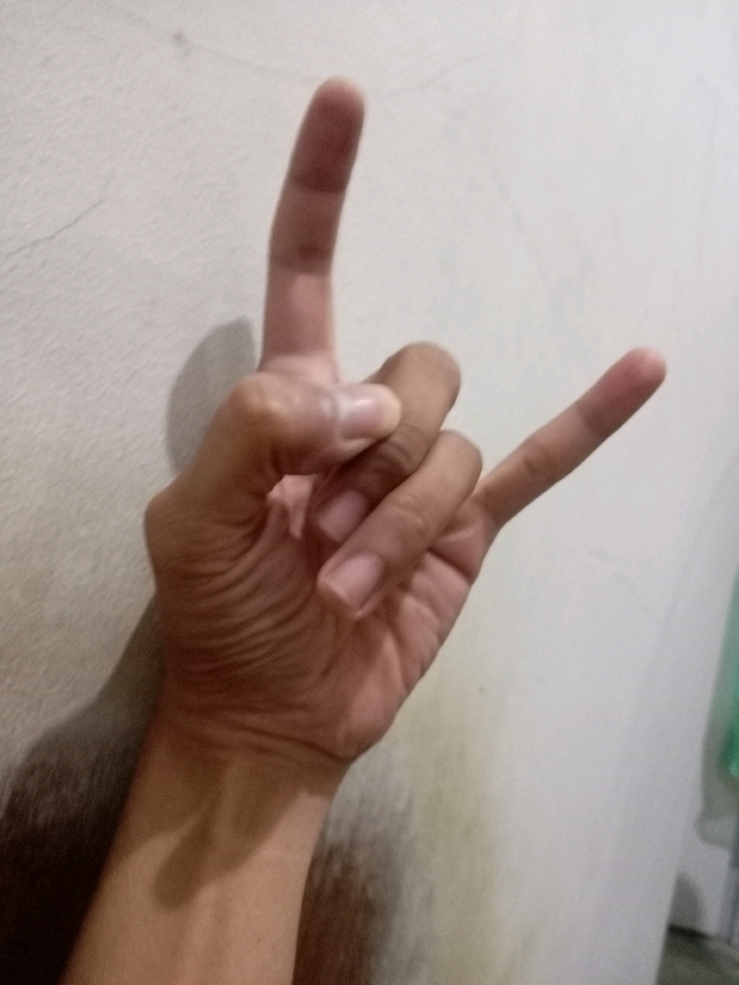 A hand is shown with the index finger and little finger extended upwards, while the middle finger and ring finger are curled inwards, with the thumb pressed against them. This gesture is commonly known as the "rock on" sign. The hand is positioned against a textured, off-white wall, casting a shadow to the left. The lighting is dim, and the image has a grainy quality. The specific location in Batam, Indonesia cannot be determined from the provided image.