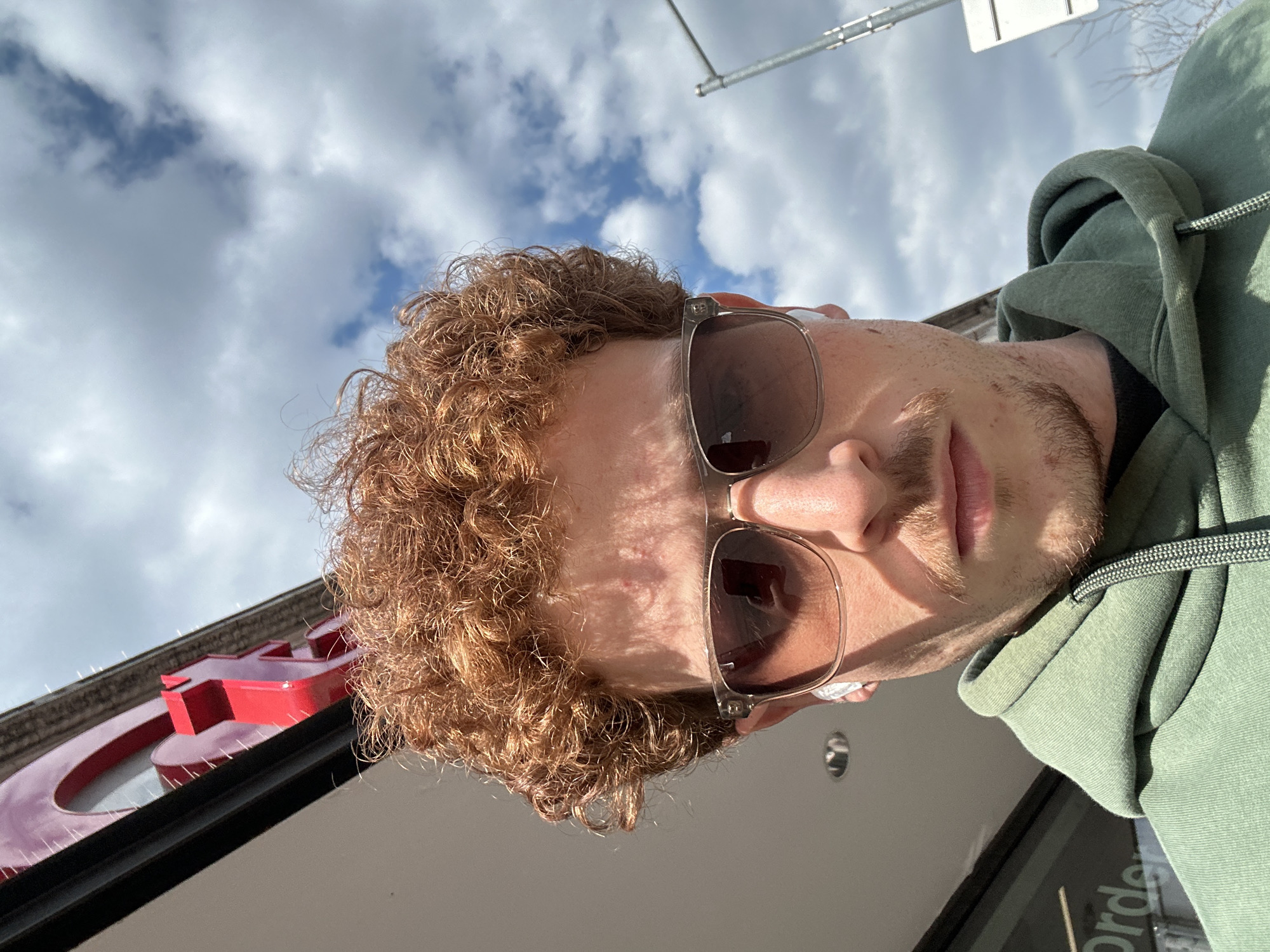 A close-up, upward-angled outdoor portrait depicts a young adult male in the city of New York, United States of America. The subject has reddish-brown curly hair, a light brown mustache, and stubble beard. He wears clear-framed sunglasses with dark lenses and a white earbud in his left ear. His attire consists of a sage green hooded sweatshirt over a dark undershirt. The background on the upper left features a red, three-dimensional letter, resembling an "A," mounted on a building facade. Below this sign, a dark horizontal building element is visible, along with faint text that appears to read "Order" on a surface. The upper right background displays a partially cloudy blue sky and a metallic grey street pole with a partial white rectangular sign attached. Bare tree branches are visible in the far distance on the right.