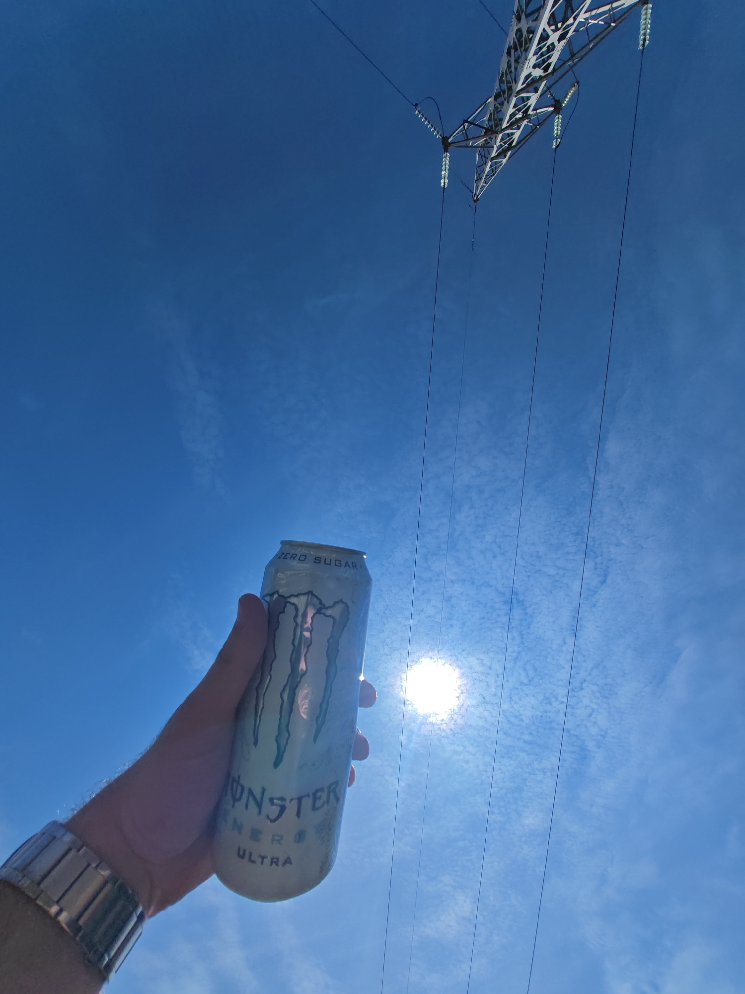 An outdoor, low-angle photograph, taken in what is presumed to be the vicinity of Shumen, Bulgaria, depicts a human hand holding a 'Monster Energy Ultra' beverage can against a bright, partly cloudy blue sky.
**People:** A human left hand, showing a metallic, link-bracelet watch on the wrist, holds the can. No other individuals are visible.
**Subjects/Objects:**
* **Beverage Can:** A vertically oriented, silver-white 'Monster Energy Ultra' can is held in the center-left foreground. The distinctive green Monster claw logo is visible on its surface, along with "MONSTER ENERGY ULTRA" text and "ZERO SUGAR" near the top rim.
* **Watch:** A metallic (likely stainless steel) watch with a segmented link bracelet is visible on the wrist of the hand holding the can.
* **Sky and Sun:** The background is a clear, vibrant blue sky with scattered, thin, wispy white clouds. A bright, unfiltered sun creates a strong glare in the lower-right quadrant, partially obscured by the can.
* **Power Infrastructure:** In the upper-right portion of the frame, a segment of a gray, lattice-structured high-voltage electrical transmission tower is visible. Multiple dark, thin power lines extend downwards from the tower, suspended by light-colored, segmented glass or ceramic insulators.
**Actions/Interactions:** A hand is actively holding the beverage can. The bright sun illuminates the scene from behind the can.
**Environment:** The setting is outdoors, characterized by open sky and the presence of significant electrical transmission infrastructure, consistent with areas on the outskirts or industrial zones, or along transmission corridors near a city like Shumen.