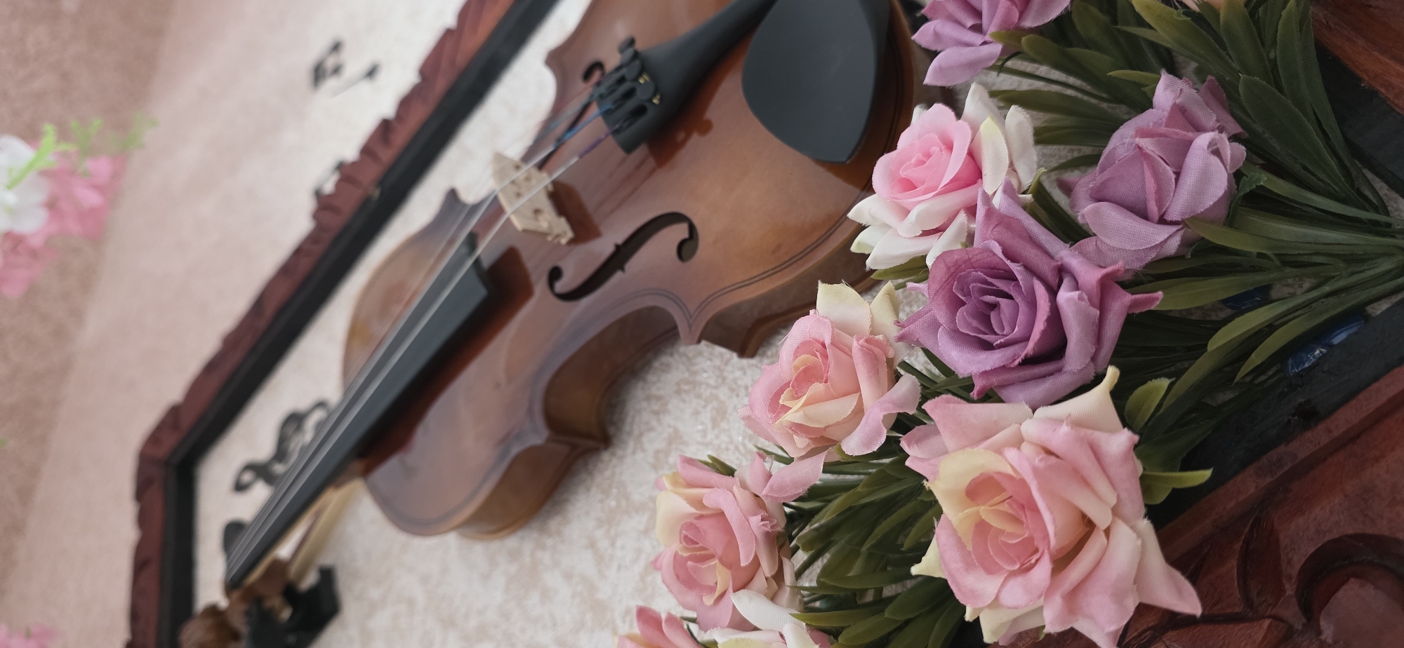 The image displays a close-up of a violin and an arrangement of artificial roses. The violin is a classic wooden instrument with a dark brown finish, resting on a textured surface. The roses are in various shades of pink and purple, with green artificial leaves. In the background, a dark-colored frame is visible. There are no people or animals present in the image. The location is not specified in the image.