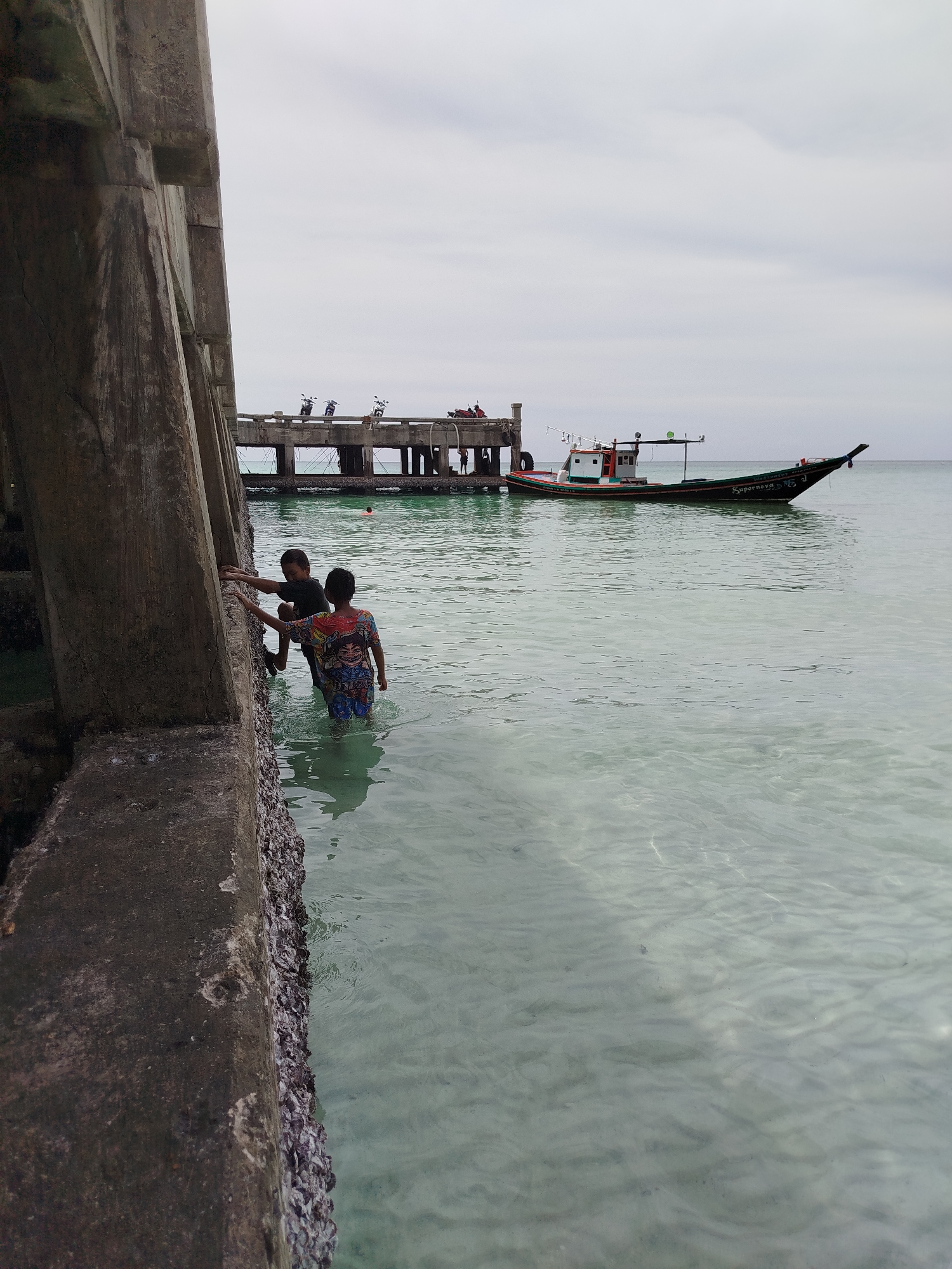 Two young boys are playing in the shallow, clear water near a concrete pier in Ko Pha-ngan District, Thailand. The boy on the left is wearing a dark t-shirt and shorts and is climbing on the barnacle-encrusted concrete structure. The boy on the right is wearing a colorful, patterned t-shirt and shorts and is standing in the water, also reaching towards the pier. In the background, a long, slender boat with a white cabin and a green hull is anchored near the pier. The pier itself is supported by concrete pillars and has several motorcycles parked on its surface. The sky is overcast, and the water is calm with gentle ripples. The scene captures a moment of childhood play against a backdrop of a working harbor.
