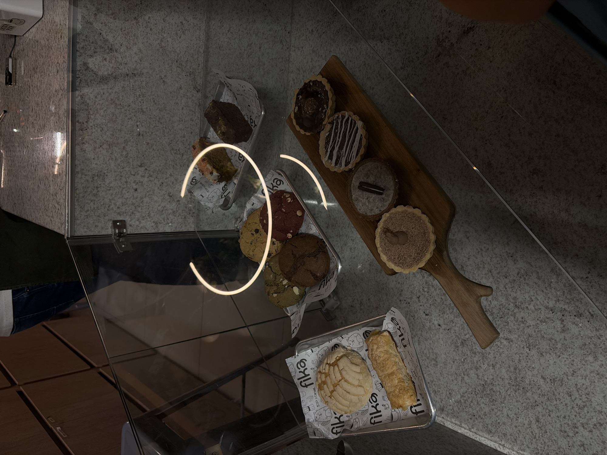 The image captures an overhead view of a diverse selection of baked goods displayed on a light-colored, speckled granite-like surface, likely within a glass display case at a bakery or cafe in Puebla, Mexico.
A prominent wooden serving board, shaped like a branch, holds four individual tarts. These include a dark chocolate tart with a chocolate confection, a tart with white icing drizzle, one topped with an Oreo cookie piece, and a light brown tart possibly flavored with cinnamon or streusel.
To the left, a metal tray lined with branded paper holds an assortment of cookies: a classic chocolate chip, a red velvet, a matcha or pistachio green cookie, and a double chocolate cookie. Above this, another metal tray contains two rectangular, dark brown baked items, resembling brownies or small loaf cakes. Below the wooden board, a third metal tray, also lined with the same branded paper, features traditional Mexican sweet bread—a concha with its characteristic shell pattern and a long, golden-brown pastry.
Reflections are visible on the glass surface, including a bright, curved neon-like light strip in the center-left. Faint reflections of people are discernible; an arm or shoulder in the top right and legs/pants in the bottom left, suggesting individuals are viewing or interacting with the display.
Visible text on the paper liners within the trays clearly shows "FIKA" in a stylized font, accompanied by smaller text "SIP & DRINK" and graphic illustrations of coffee cups and pastries. A partial view of a box in the top left corner displays "CHECK HERE". The presence of the concha and the overall presentation suggest a modern establishment offering both international and local treats.