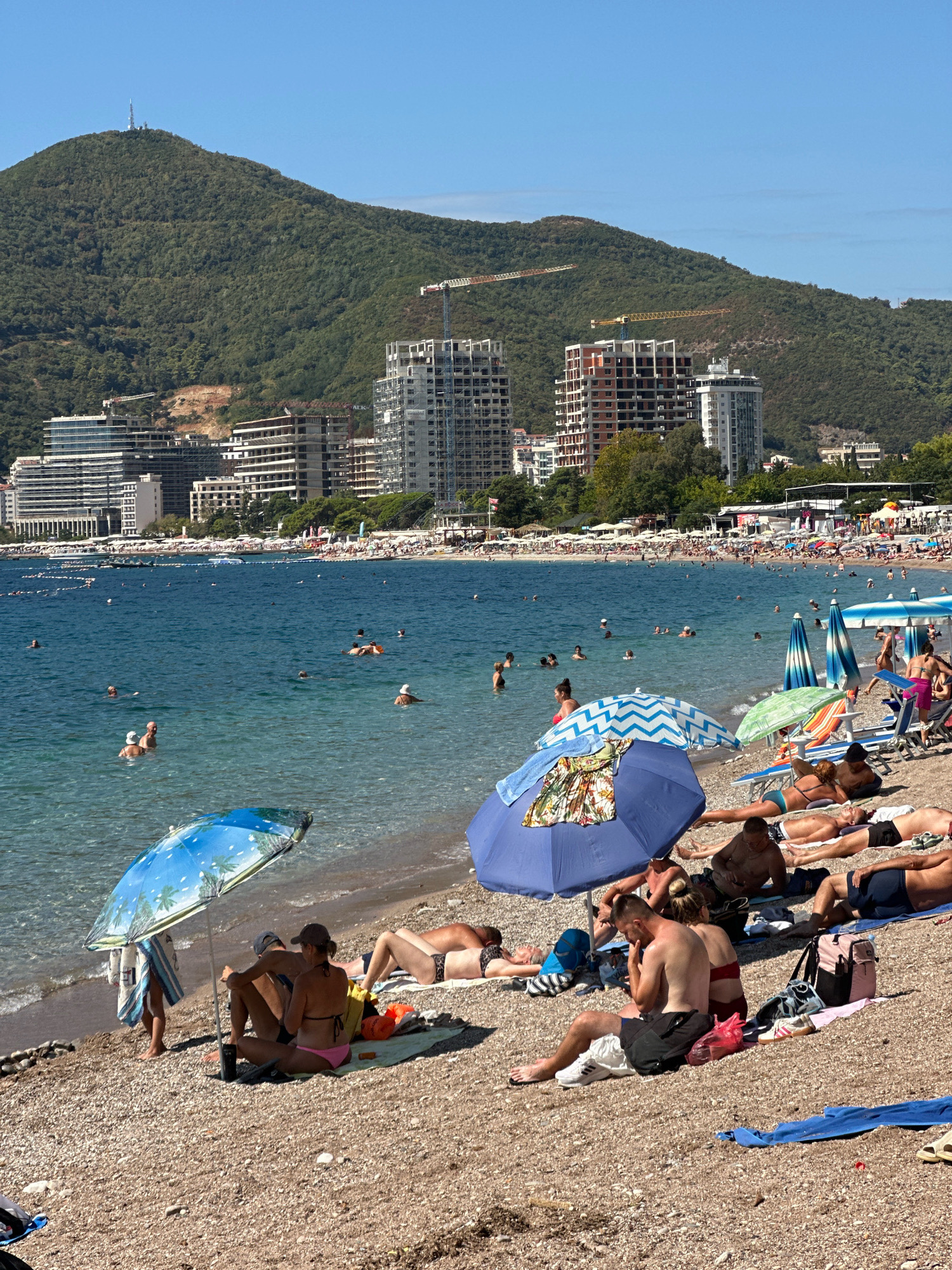 The image depicts a crowded beach scene in Budva, Montenegro, with the Adriatic Sea in the foreground and a densely built-up coastline backed by a verdant, tree-covered mountain under a clear blue sky.
On the pebbled beach, numerous individuals are sunbathing on towels and lounge chairs, while others are partially shaded by colorful umbrellas. Some people are engaged in conversation, while others are looking at their phones. A few individuals are seated under umbrellas.
In the shallow waters of the sea, several people are swimming and wading. The water is a clear, vibrant blue. Along the coastline, a long stretch of beach chairs and umbrellas indicates a popular resort area. In the background, a dense collection of modern buildings, including hotels and apartment complexes, lines the shore. Several construction cranes are visible among the buildings, suggesting ongoing development.
The mountain dominating the background is lush with vegetation and has a telecommunications tower at its summit. The overall atmosphere is one of a busy summer day at a popular coastal tourist destination.