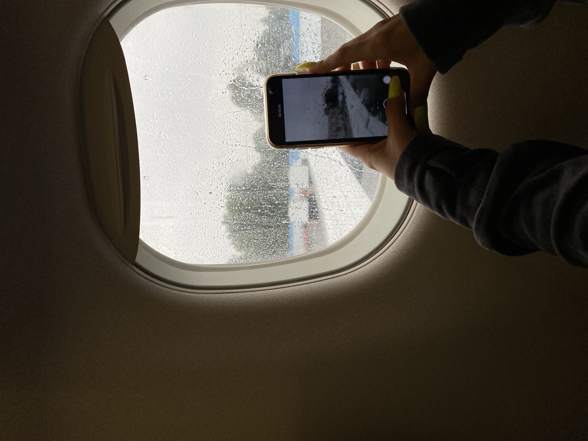 A person's hands, adorned with bright yellow nail polish, hold a smartphone in landscape orientation to capture a video of a rainy day viewed through an airplane window. The window is streaked with numerous raindrops, obscuring the view of the tarmac and trees outside. The interior of the airplane cabin, likely the wall or seat, is visible in the foreground and to the left of the window. The phone screen displays a timer counting "00:00.00", indicating that a video recording is in progress or has just started. The person is wearing a dark-colored long-sleeved shirt. The overall scene suggests a travel experience during inclement weather.