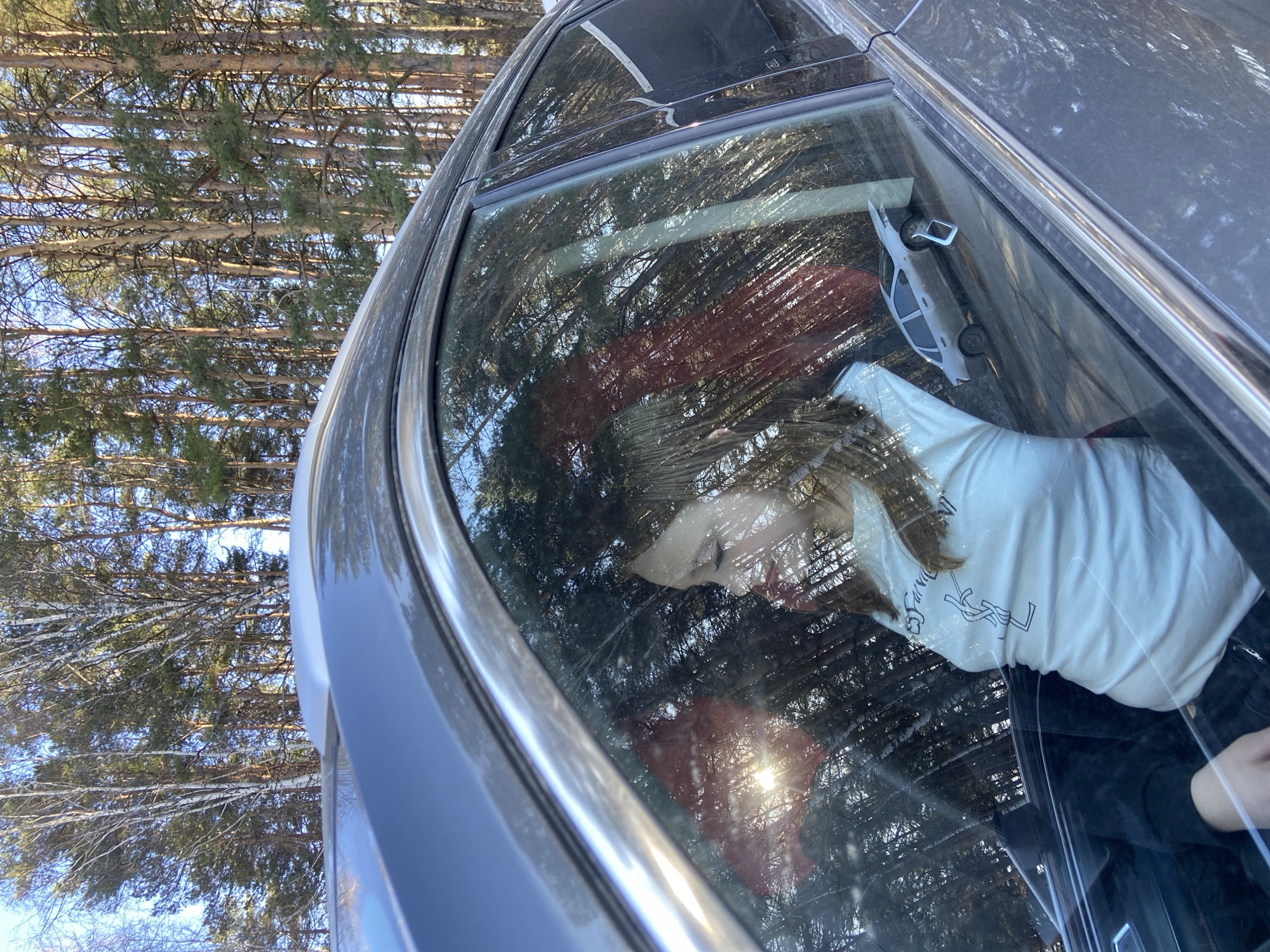 The image displays a partial view of a dark-colored car, likely an SUV or sedan, from its side and roof, parked outdoors in a forested area. A person, appearing to be female, is seated inside the car, visible through the passenger window. They are wearing a white short-sleeved t-shirt featuring a prominent black logo resembling the interlocking "YSL" monogram. Dark pants are also visible.
Inside the vehicle, a small white toy car or model is placed on the dashboard. A red fabric, possibly a blanket, is visible in the back seat, and a white object or bag is seen near the person's lap. The car windows show strong reflections of the surrounding environment and bright sunlight.
The background consists of numerous tall, slender trees, a mix of coniferous (pine/spruce) and deciduous types, including some with distinct white bark characteristic of birches, many of which appear bare. The sky is clear and blue, indicating a sunny day. Traces of what might be snow or frost are visible on some branches, suggesting a colder season. The overall scene depicts a car parked in a wooded environment, consistent with a location like Chaikovsky, Russia. The person appears to be stationary within the vehicle.