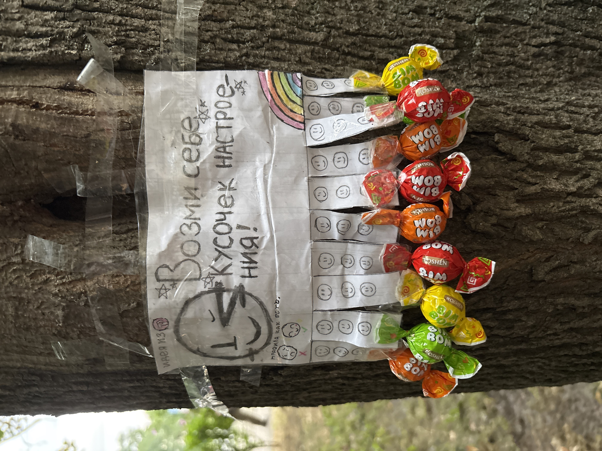 The image shows a piece of paper taped to a tree trunk. The paper has handwritten text and drawings. At the top, it says "Возьми себе кусочек настроения!" which translates to "Take a piece of mood for yourself!". Below this text, there is a drawing of a smiley face. Underneath the smiley face, there are drawings of two sad faces with red X's and two happy faces with green checkmarks, with the text "правила как есть" meaning "rules as they are". Below this, there are columns of perforated tear-off strips, each with a drawing of a smiley face. At the bottom of the paper, there are multiple colorful candies attached with tape, some of them appearing to be from the brand "BOM-BOM". The tree trunk is textured and appears to be outdoors, with some green foliage and branches visible in the background, suggesting a park or a street setting. The overall scene appears to be an act of kindness or a community art project, offering small treats and a positive message. The context of Odesa, Ukraine, is not directly discernible from the visual elements of the image itself, but can be inferred as a known location for such initiatives.