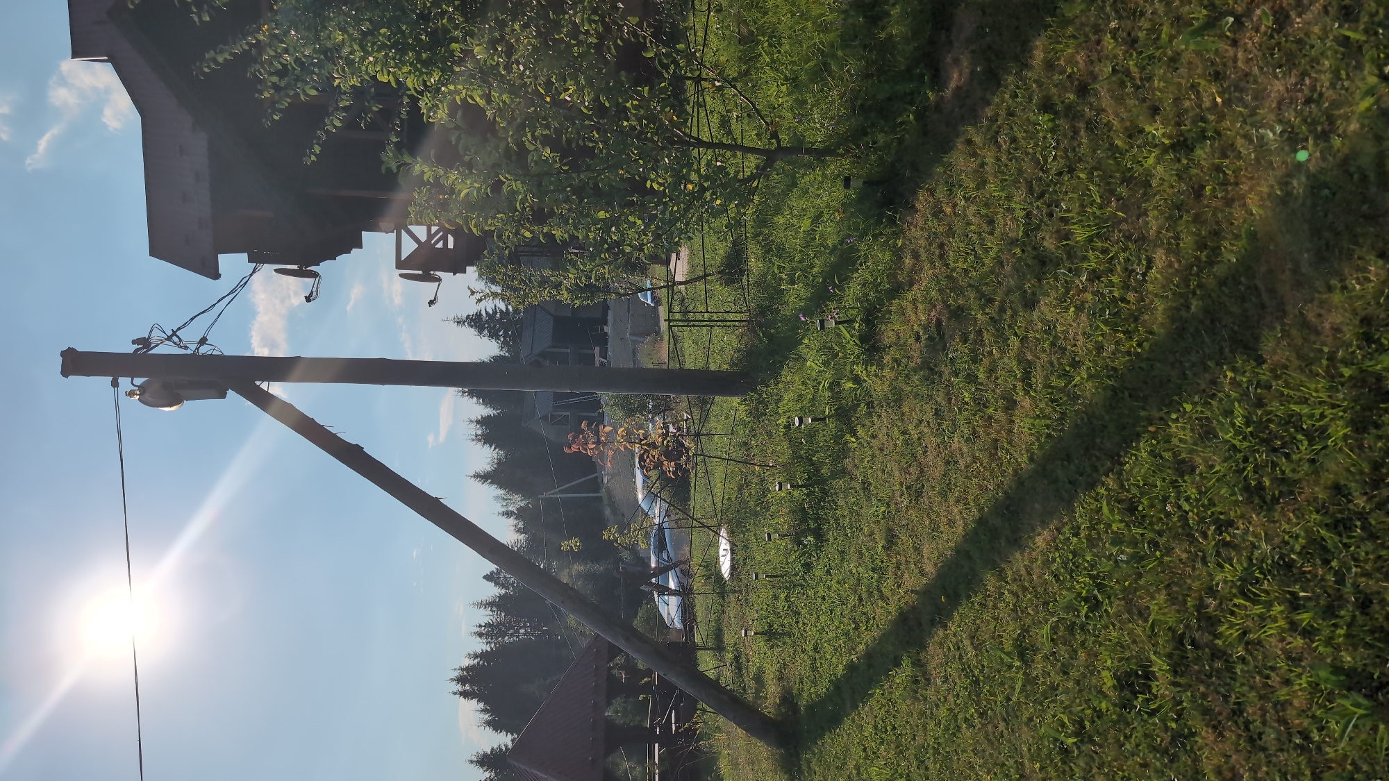 The image depicts an outdoor scene in Yablunytsya, Ukraine, likely during daylight hours given the strong sunlight. The foreground is dominated by a grassy lawn with scattered small, dark-colored garden lights. A long shadow, cast by an unseen object, stretches diagonally across the lawn from the upper left to the lower right.
In the midground, a wooden utility pole with a diagonal support pole stands prominently. Wires are suspended from the top of the pole, connecting to equipment that appears to be a transformer. To the right of the utility pole, a portion of a wooden building with a dark roof is visible. Two satellite dishes are mounted on the side of this building.
Behind the utility pole and extending to the left, a black metal fence separates different areas of the property. Through the fence, several vehicles are parked, including what appears to be a silver car. Further in the background, a dense line of evergreen trees forms a natural boundary. Beyond the trees, additional wooden structures, possibly homes or other buildings, can be seen on a hillside, suggesting a semi-rural or mountainous setting. A small gazebo with a dark, multi-sided roof is visible in the far left of the midground. A young tree with reddish-orange leaves is situated in front of the fence. The sky above is blue with some white clouds.