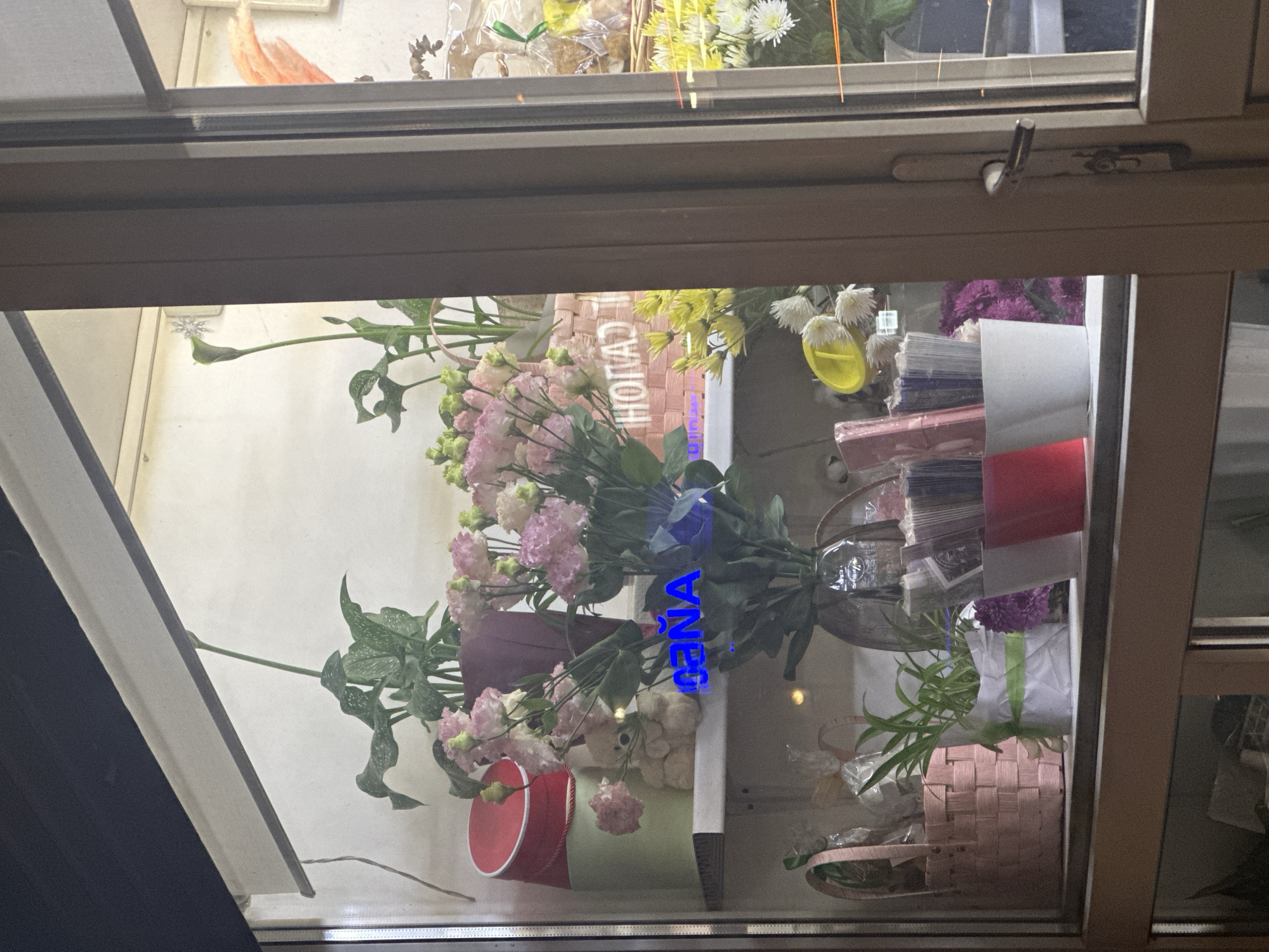 The image captures a vibrant display of flowers and related merchandise, likely viewed through a shop window or a display case. The scene is brightly lit, showcasing various floral arrangements and accompanying items.
The main subjects are bouquets of fresh flowers. Prominently featured are delicate pink and white lisianthus, and cheerful yellow and white chrysanthemums, interspersed with green foliage. These flowers are arranged in an assortment of containers, including a clear glass vase, a light pink woven basket, a red cylindrical box, and a green cylindrical pot.
Among the flowers, other objects for sale or display are visible: stacks of colorful wrapping materials (red, white, and purple/blue paper or cardstock), a small yellow scoop or watering can, and a fluffy white decorative item. The background behind the display appears to be a plain, light-colored wall.
A notable detail is the prominent blue illuminated text, possibly a neon sign or reflection on the glass, which clearly spells out "ЦЕНА" (CENA), meaning "PRICE" in Russian. This text, along with the nature of the display, strongly indicates a commercial setting, specifically a flower shop in a Russian-speaking region like Stavropol, Russia.
The image shows a static presentation of goods; no people or specific activities are depicted. The foreground includes parts of a dark window frame and a window latch, suggesting the photo was taken from outside, looking into the shop display. The time of day appears to be during operating hours, with ample artificial lighting illuminating the display.