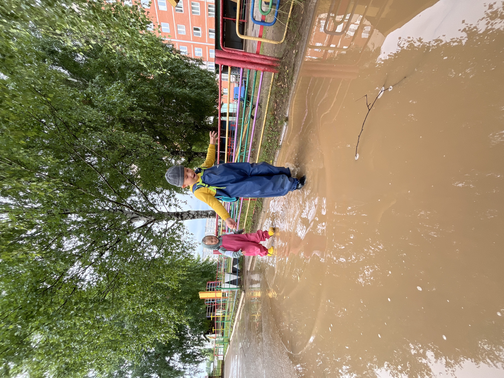 In Chaikovsky, Russia, after a rain, two young children are enjoying a large puddle in a courtyard. A boy in a yellow shirt and blue overalls is walking through the muddy water, splashing with his feet. Beside him, a smaller child in a pink jumpsuit and yellow boots, holding the boy's hand, is also navigating the puddle. The background shows a playground with colorful fencing, trees with lush green leaves, and a residential building. The sky is overcast, indicating recent or ongoing rain.