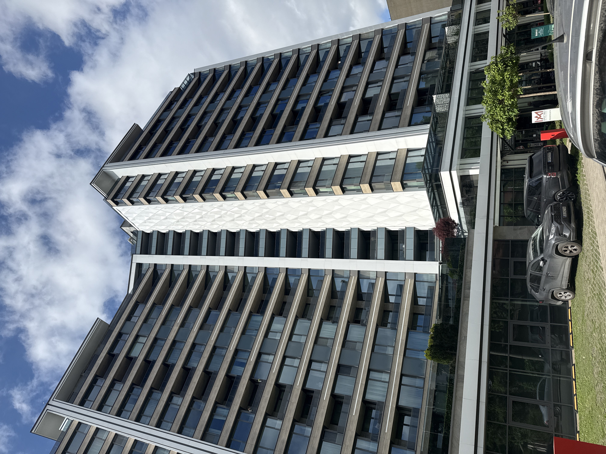 The image depicts a tall, modern building with a unique architectural design. The building has numerous windows and is constructed with a combination of materials, including glass, concrete, and metal. The building appears to be located in an urban area, likely a city, as evidenced by the presence of vehicles and street infrastructure nearby. The sky is partially visible with a mix of blue sky and white clouds. The foreground of the image shows two cars parked along the side of the building. The image is taken from a low angle, looking up at the building, which emphasizes its height and grandeur.
