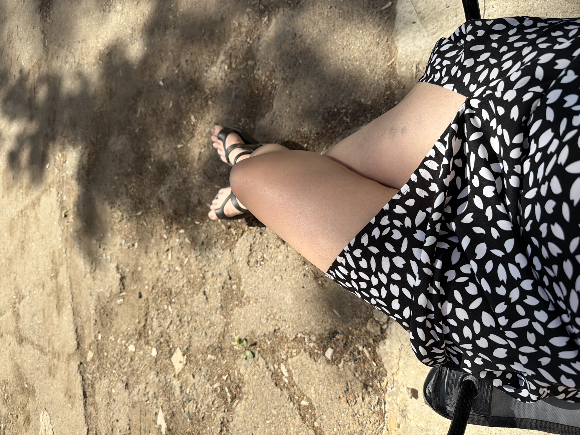 A high-angle shot depicts a person sitting in a chair on a dirt or gravel surface, their legs crossed at the knees. The person wears black sandals and a black and white patterned dress that drapes to their right. The ground is partly shaded by a tree. The city and location are unknown.