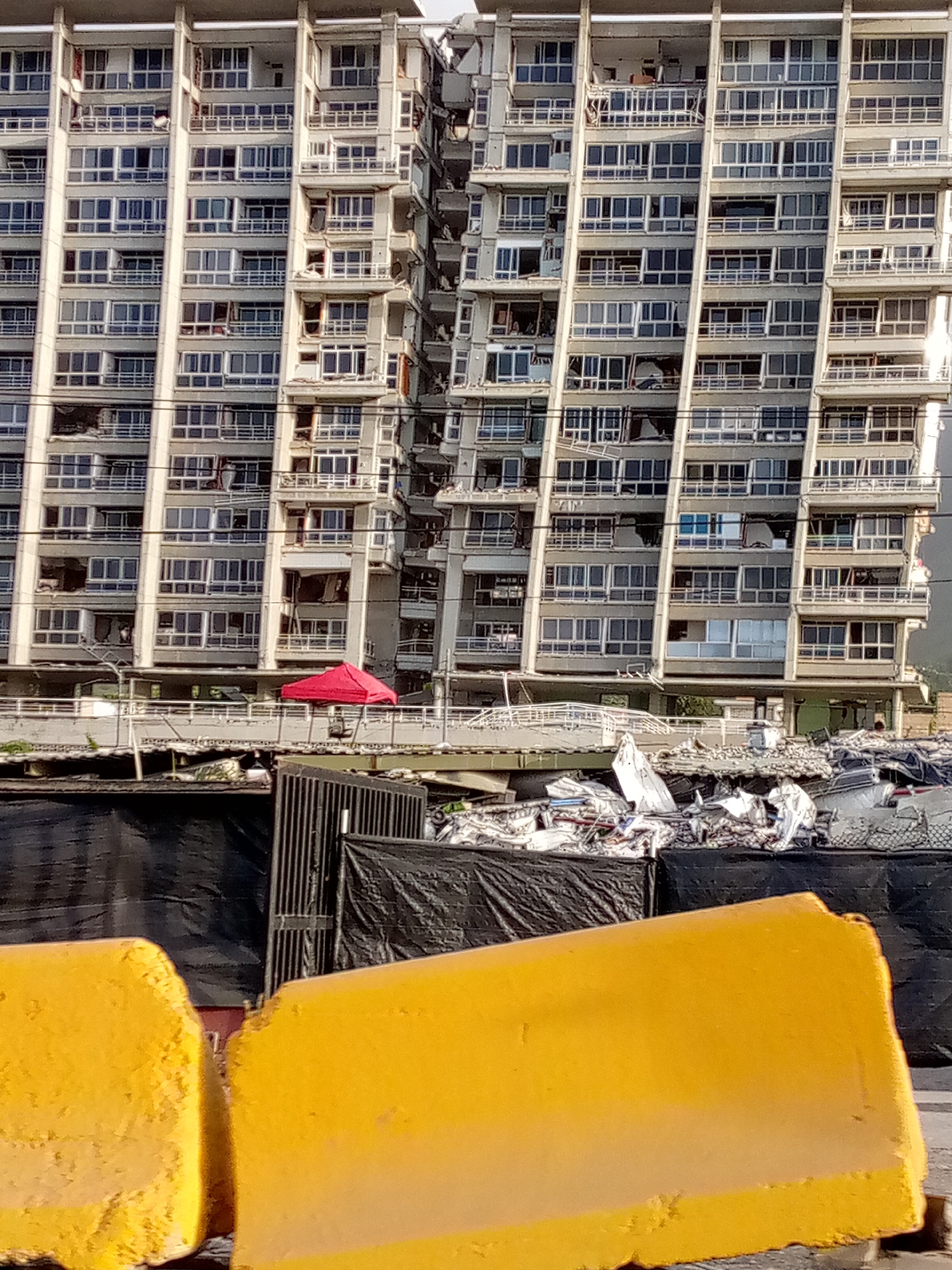 The image shows a severely damaged apartment building in Caraballeda, Venezuela, likely after a natural disaster. The building's facade displays numerous broken windows, collapsed balconies, and structural damage, indicating significant destruction. In the foreground, yellow construction barriers are visible, along with black tarps and debris piles, suggesting ongoing cleanup or recovery efforts. A small red tent is pitched on a platform at the base of the building, possibly providing temporary shelter or a staging area for rescue workers. The scene is one of devastation and the aftermath of a catastrophe.