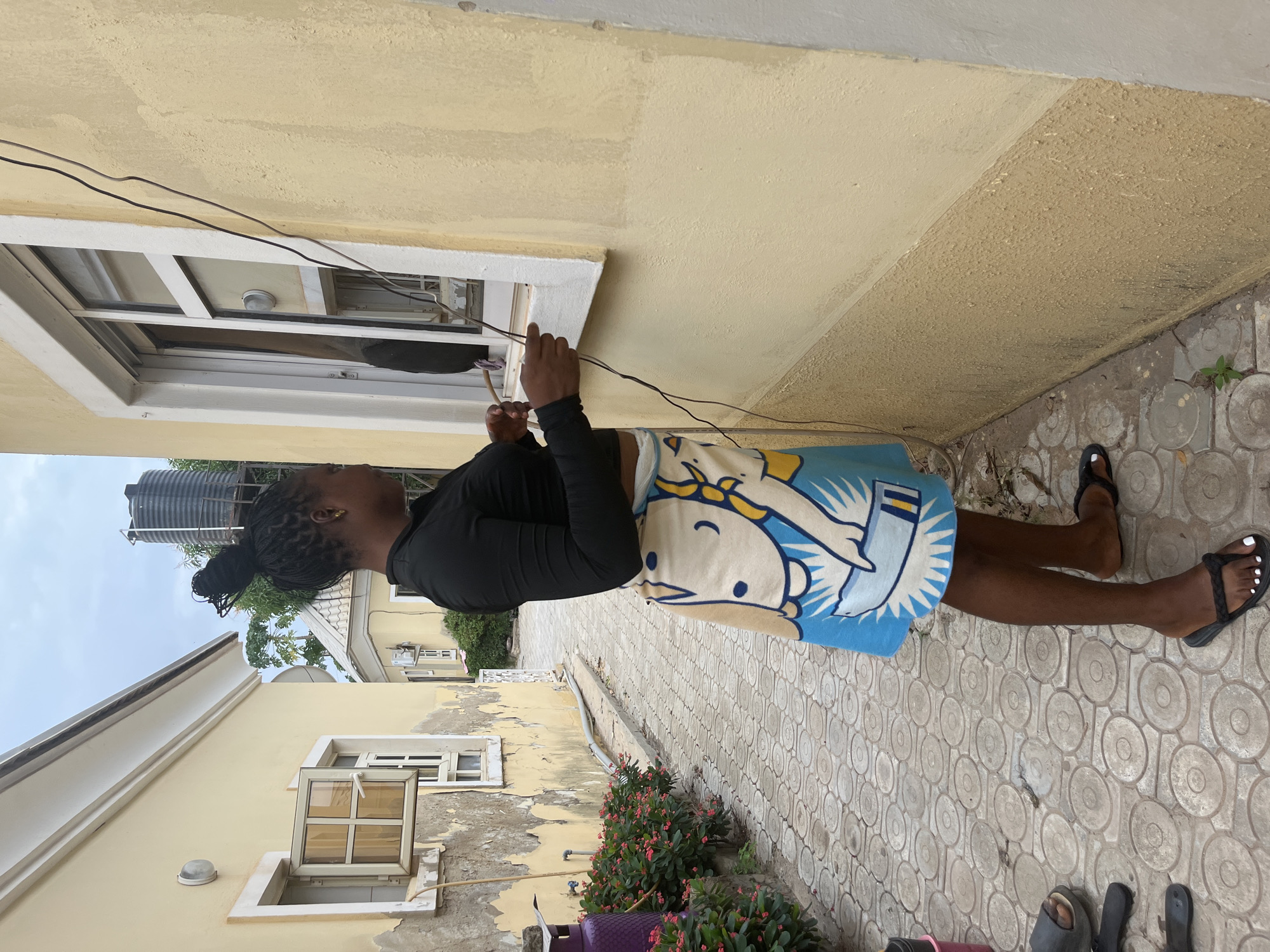 In an outdoor alleyway in Abuja, Nigeria, a Black woman with her hair styled in a bun, wearing a black long-sleeved shirt and a patterned skirt, stands facing a light yellow building. She is positioned with her left side towards the viewer, looking towards a window of the building and holding a thin object, possibly a stick or wire. The building's facade is textured and weathered, with white trim around the window. Electrical wires run along the exterior wall. The ground is paved with circular stone patterns. To the left of the woman, there are green bushes with pink flowers and a purple water dispenser. In the background, another building with a water tank on its roof is visible. The sky above is overcast.