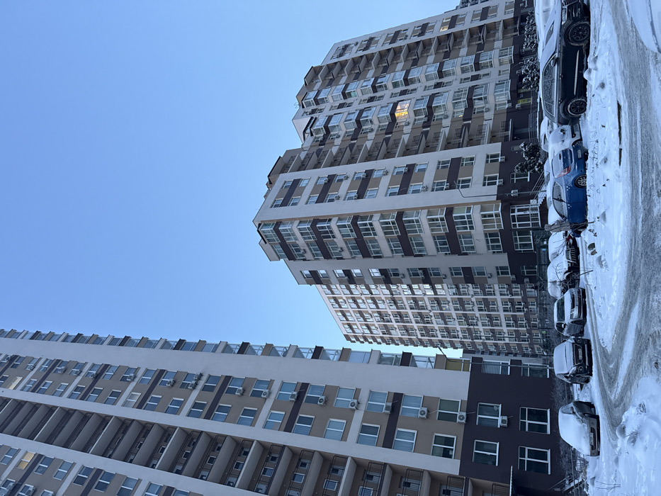 Two tall apartment buildings stretch skyward against a pale blue sky. The buildings feature repetitive window patterns with white and beige facades. Parked cars line the street along the buildings' sides, partially covered in snow. The image is taken from a low angle, emphasizing the buildings' height.