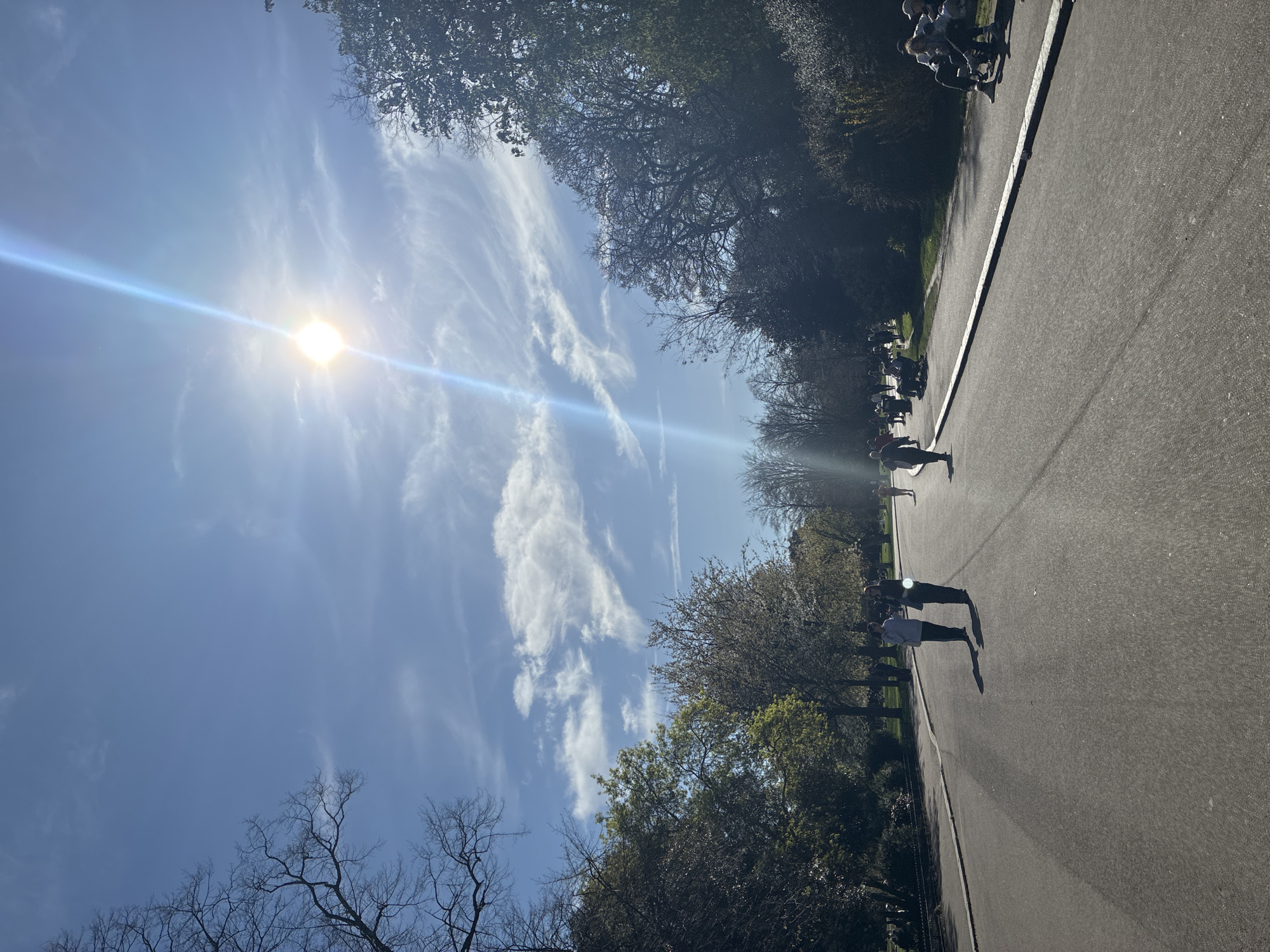 A ground-level, long shot in a London park during daylight hours shows a wide asphalt path running diagonally through the frame, bounded on either side by trees and shrubbery. The sky above is predominantly blue, with scattered white clouds and a bright sun creating a lens flare. People are walking along the path.