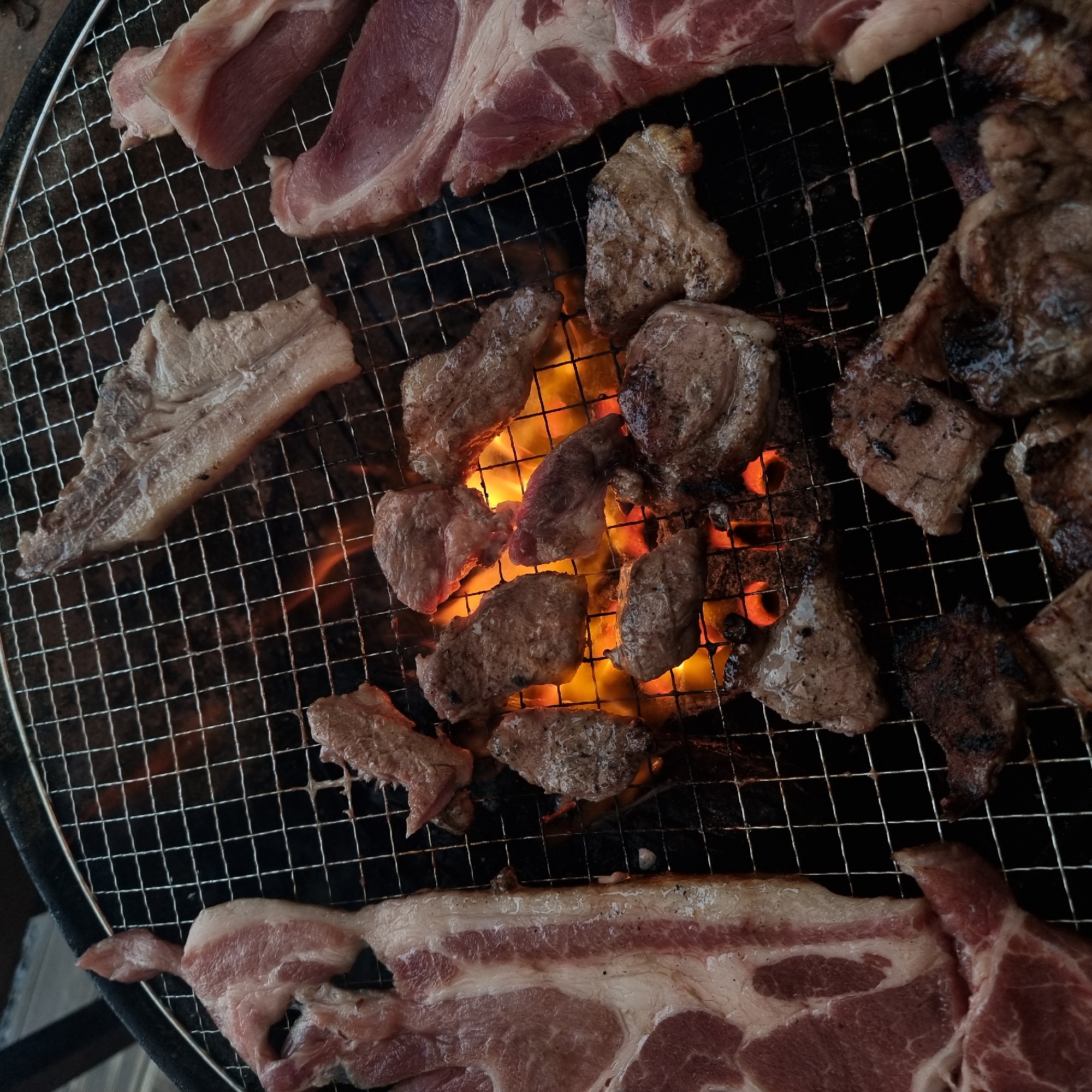 This is a close-up, overhead shot of various cuts of raw and partially cooked meat grilling over hot coals. The scene appears to be outdoors, on a metal grill grate, with flames visible beneath. The meat includes thick slices with visible fat marbling and smaller, more uniformly shaped pieces. The grill grate is a grid of metal wires. The background is dark, suggesting nighttime or a shaded area. The overall impression is of a barbecue or outdoor cooking event. The image does not contain any discernible information about the specific location within Dunnae-myeon, South Korea.