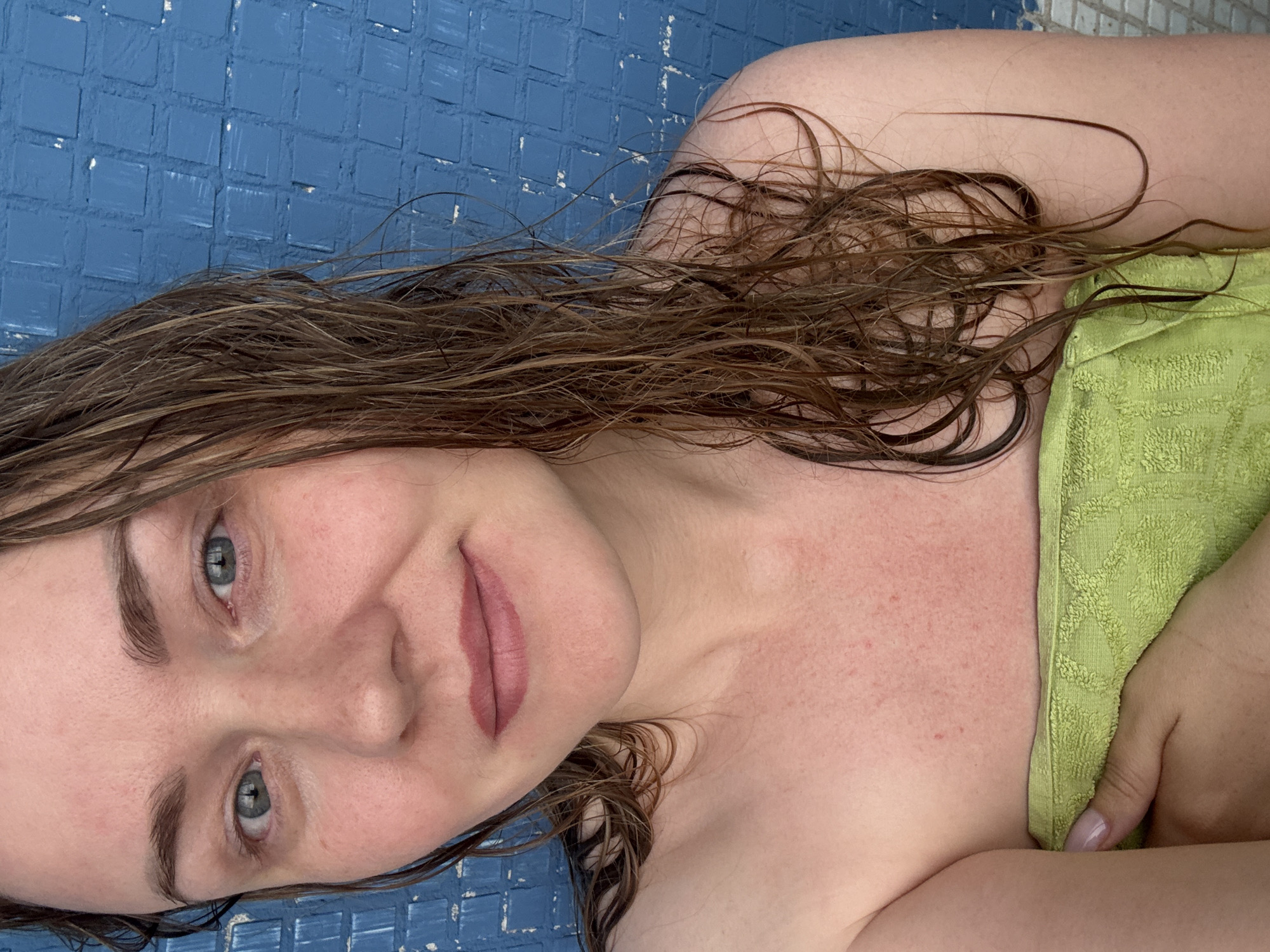 A medium shot shows a fair-skinned woman with wet, brown hair wrapped in a light green towel, taken from the chest up. She has light blue eyes, full lips with a pink tint, and a smattering of freckles across her nose and cheeks. Her eyebrows are dark and well-defined. She appears to be smiling subtly. Behind her, a blue tiled wall serves as the backdrop. The lighting suggests an indoor setting, possibly a bathroom or near a pool.
