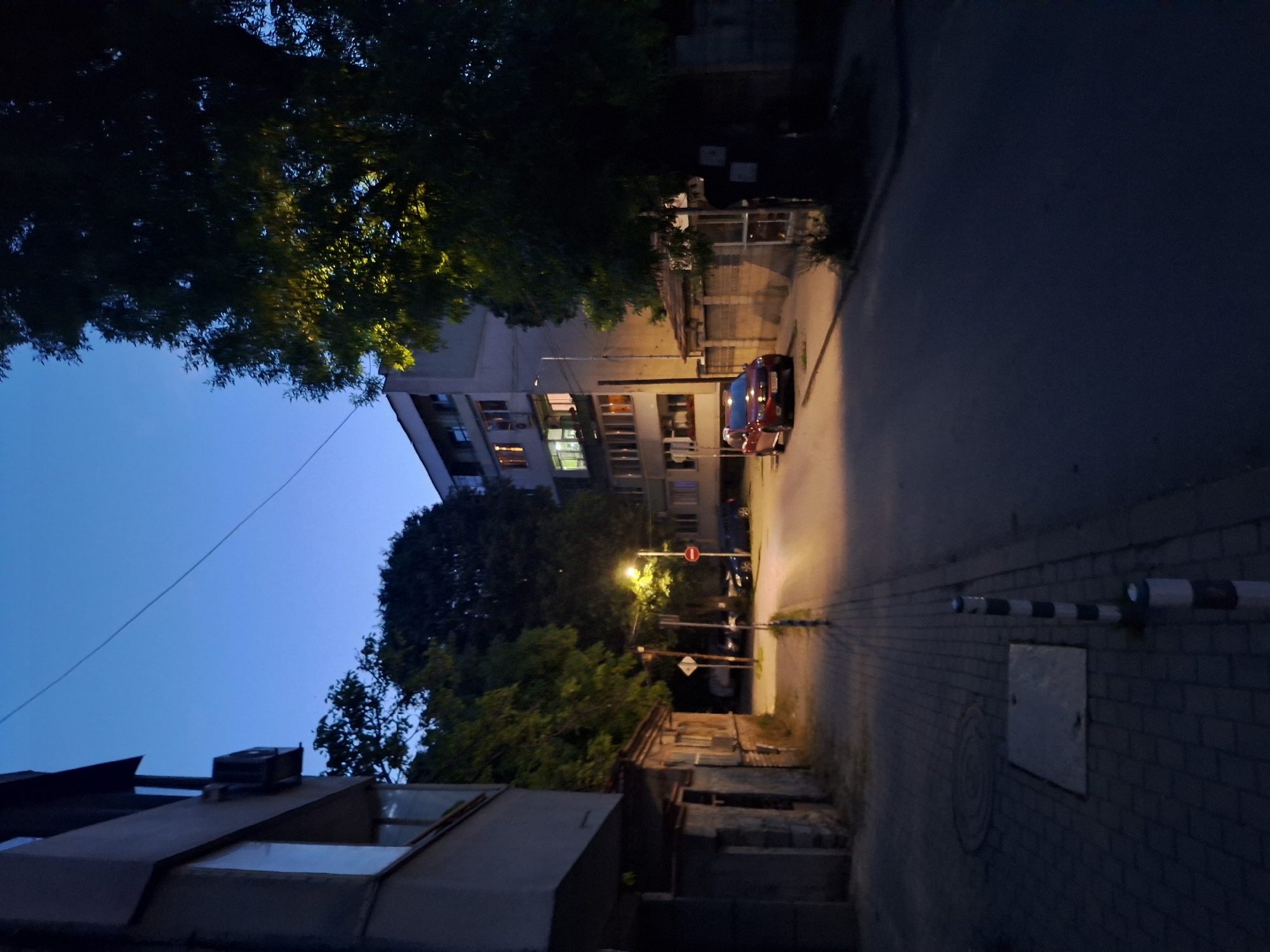 The image depicts a dimly lit street scene in the city of Varna, Bulgaria, during twilight. On the left, a modern apartment building with large windows and an air conditioning unit is visible. Adjacent to it is a low-lying, older stone building with several windows. The street is paved with cobblestones on the sidewalk and asphalt on the road. A street lamp illuminates a portion of the road and sidewalk.
A red sedan is parked on the right side of the street. Further down the road, another dark-colored vehicle is parked. There are also traffic signs present, including a red circular sign with a diagonal bar. Large trees with dark foliage line the right side of the street, with one tree's canopy illuminated by the street light. The sky is a deep blue, characteristic of early evening. The overall impression is one of a quiet urban street as night begins to fall.