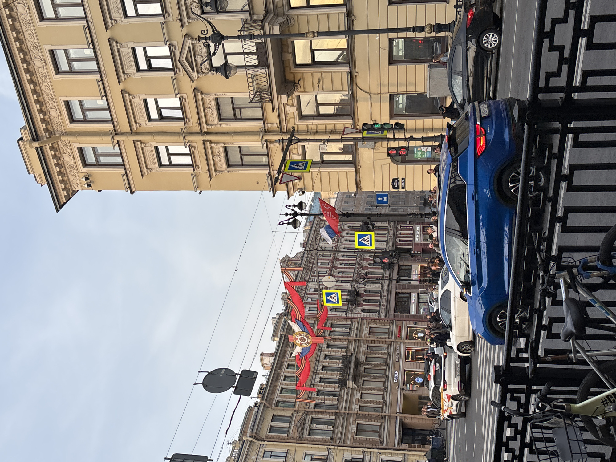 The image shows a street scene in Saint Petersburg, Russia, on a clear day. Buildings line both sides of the street, which is filled with parked cars and pedestrian traffic. The architecture is ornate, with detailed facades and multiple windows. Red ribbons and Russian flags are displayed across the buildings, likely decorations for a holiday or celebration. There are pedestrian crossing signs hanging above the street. On the right side of the image, a black metal fence and bicycles are visible. People are walking along the sidewalks and observing the scenery.