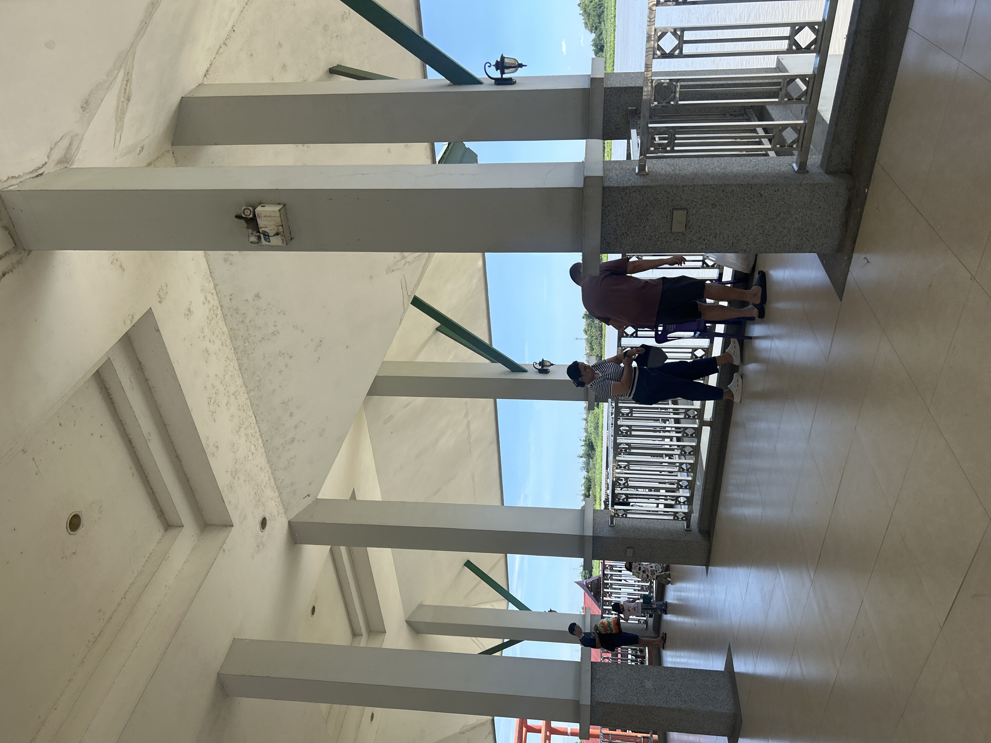 The image shows a covered outdoor walkway with multiple concrete pillars supporting a white roof. The walkway has a tiled floor and a railing overlooking a body of water. Several people are visible in the walkway, some standing and others with children. The environment suggests a waterfront area, possibly a pier or a riverside promenade. The sky is clear and blue. The location is specified as Thai Ko Subdistrict, Thailand.