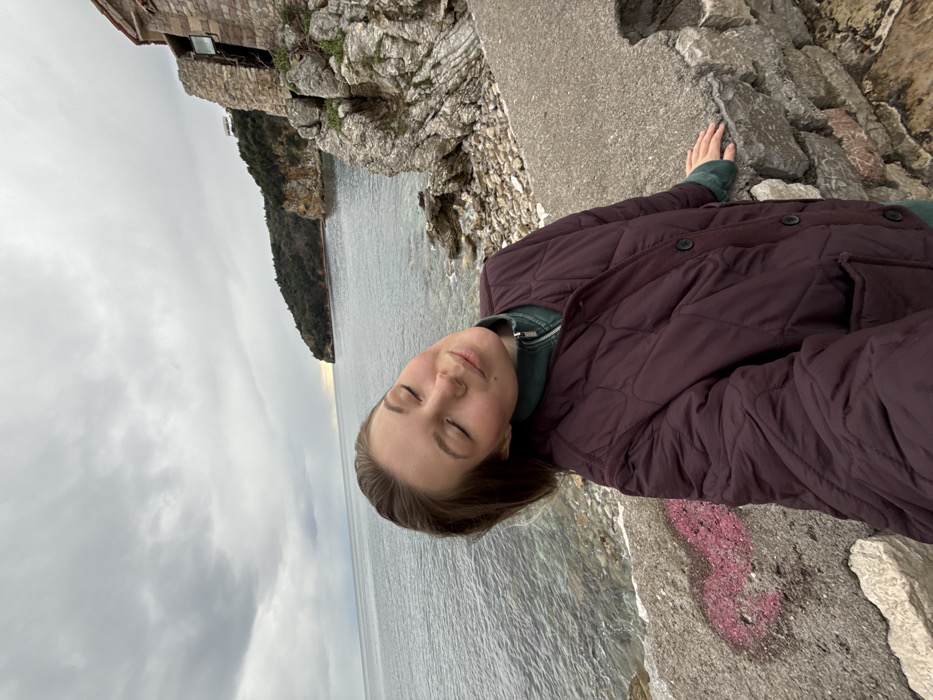 The image shows a female subject in a coastal environment adjacent to Budva Old Town, Montenegro, specifically along a path overlooking the Adriatic Sea. The woman, with light brown hair, is positioned slightly left of center, appearing to be taking a selfie. She wears a dark reddish-brown quilted jacket over a dark green zippered top. Her eyes are closed, head tilted slightly left, and she displays a subtle smile. Her left hand rests on a weathered concrete and stone retaining wall in the foreground, which bears faint pink graffiti on its concrete section. To the right, a multi-tiered stone structure, possibly part of Budva's Old Town fortifications, is integrated into a rocky cliff face, featuring visible window openings. The middle ground consists of calm, blue-green sea water with a rocky shoreline. A vegetated, rocky headland is visible in the distance to the right, beyond which a strip of brighter light, consistent with sunrise or sunset, appears on the horizon under an overcast, cloudy sky.