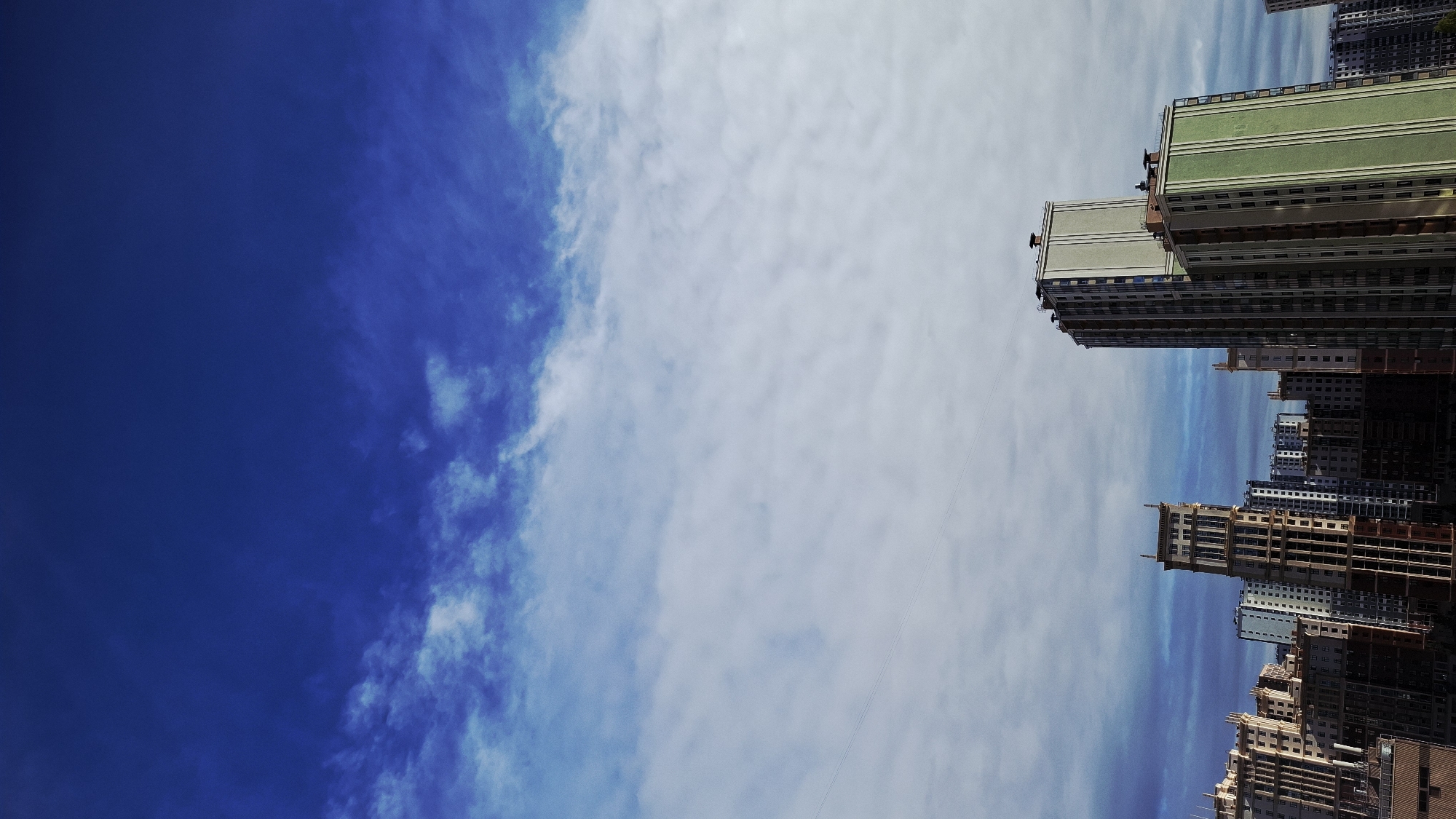 The image is an upward, angled view of the sky and buildings in Saint Petersburg, Russia. The left side of the image is mostly a deep blue sky with faint clouds, transitioning to a cloudier, lighter blue and white sky on the right. Along the right edge of the image, a cluster of tall buildings is visible, appearing to be high-rise residential or commercial structures. The buildings vary in color, some are beige or light brown with vertical lines, while others are shades of green and grey. The perspective suggests the photo was taken from a lower vantage point, possibly street level. The buildings are densely packed and appear to be under construction.