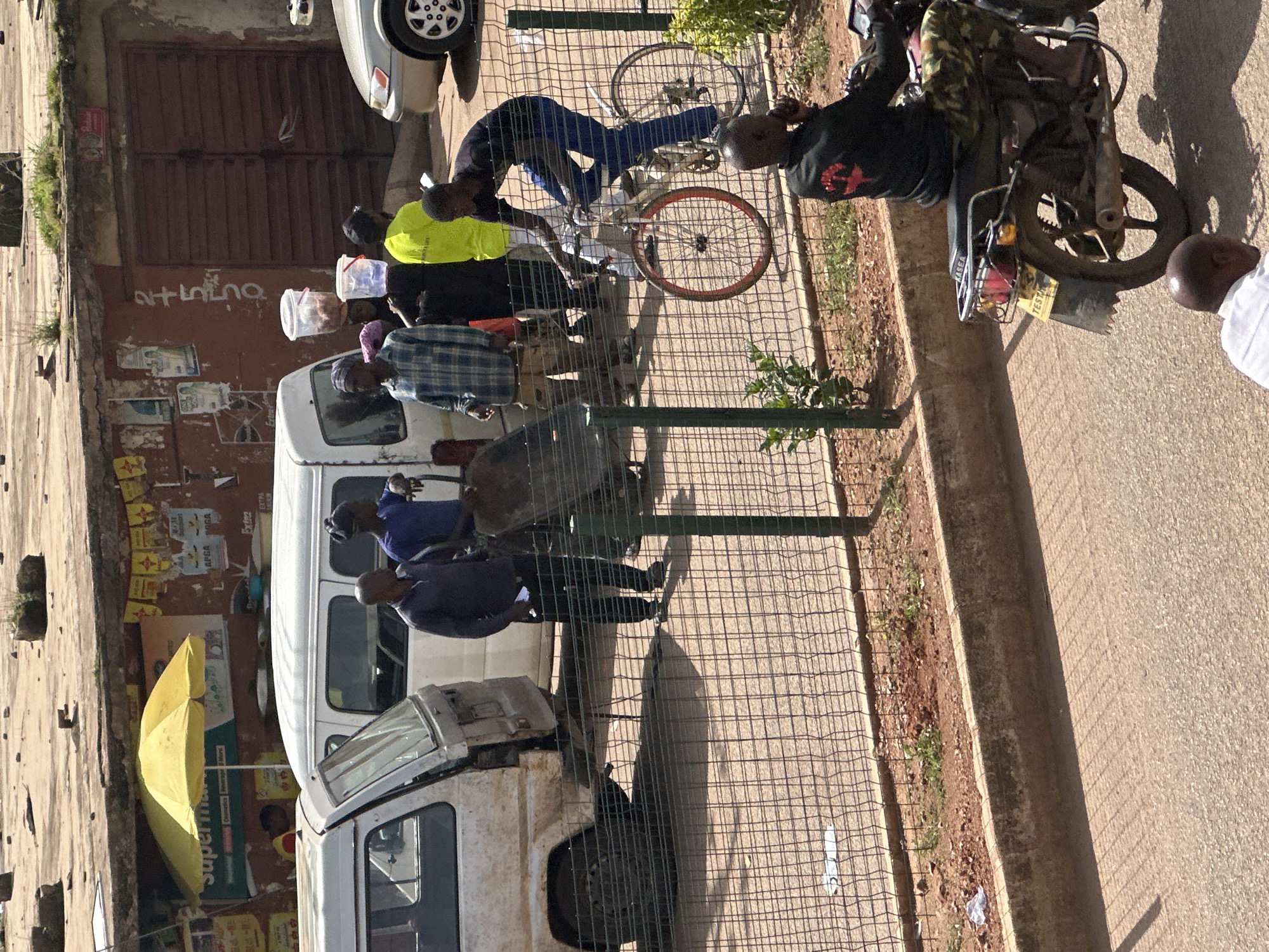The image captures a street scene in Bukuru, Nigeria, likely during the daytime given the bright sunlight casting distinct shadows. The setting appears to be an urban area, with a building facade featuring weathered walls and a large, corrugated metal door. Several people are present, engaged in various activities.
In the foreground, a man in a black shirt with a red graphic on the back is on a motorcycle. He is wearing camouflage-patterned trousers and appears to be interacting with the motorcycle. Another individual, wearing a white shirt, stands nearby, partially visible.
Further back, a group of people are gathered near a white van. Some are looking at something on the ground, possibly interacting with a bicycle or other objects. One person wears a bright yellow vest, and another is dressed in a blue shirt. A yellow umbrella is open and leaning against the building. The scene is bordered by a wire fence, and a concrete curb separates the paved ground from a patch of dirt with some sparse vegetation.
The overall atmosphere seems active and dynamic, suggesting daily life and commerce. The buildings and vehicles appear well-used, indicating a lived-in urban environment. There is no obvious indication of specific events, but rather a snapshot of everyday activity on a street. Some posters or advertisements are affixed to the building's walls, though the text is largely illegible. The presence of multiple people and vehicles suggests a relatively busy street.