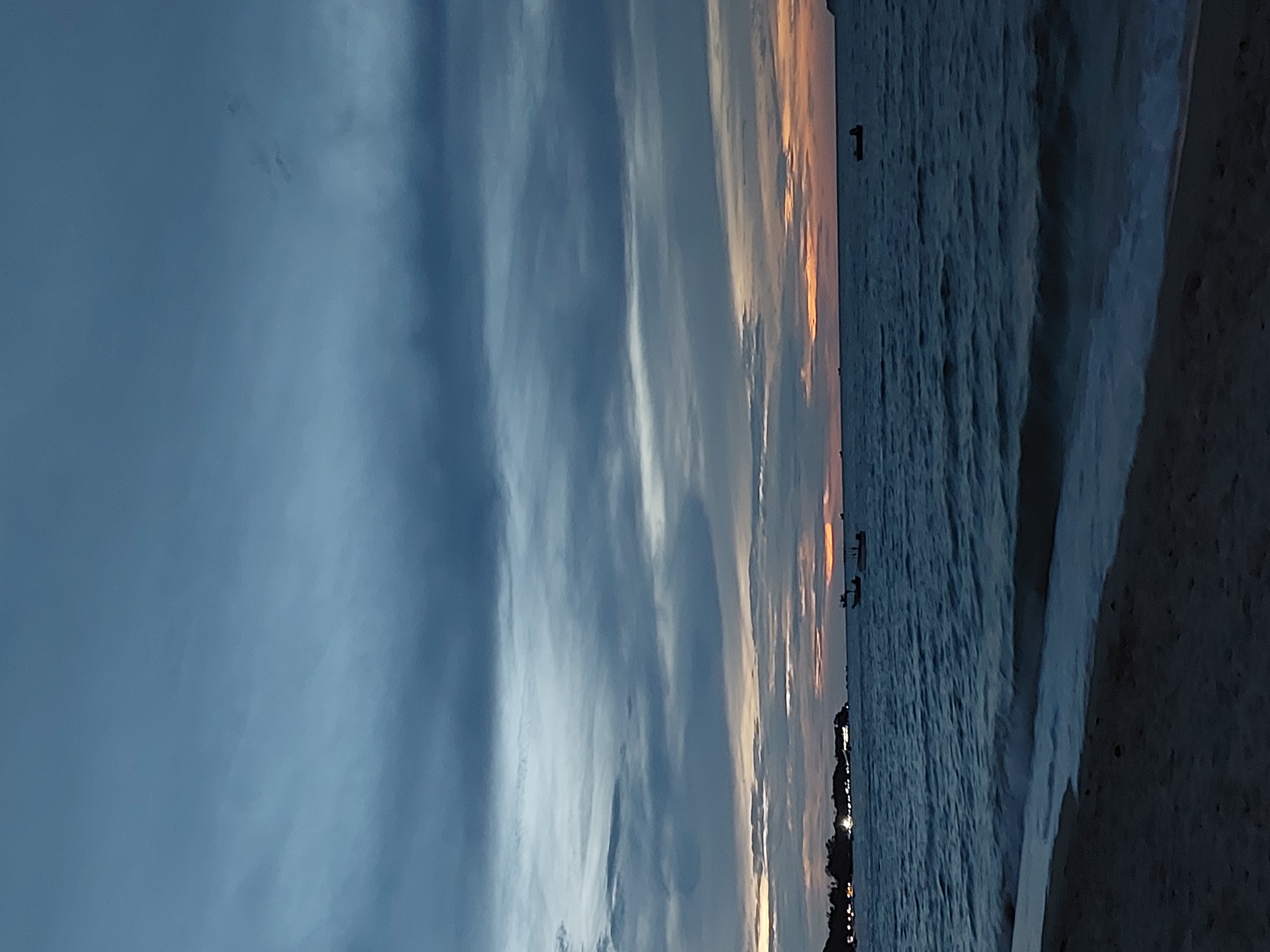This media file depicts a serene coastal scene at sunset in Baan Mae Nam, Thailand. The image is oriented vertically, showcasing a wide expanse of ocean meeting a sandy shore. The sky is a dramatic canvas of blues, grays, and oranges, with clouds streaked across, reflecting the fading sunlight. The ocean itself is a deep blue, with gentle waves lapping against the shore. A subtle glow emanates from a distant shoreline, hinting at a settlement or town.
Several small boats are visible on the water, suggesting fishing or leisure activities. One boat in particular, closer to the shore, is clearly identifiable. The presence of these boats, along with the faint lights on the horizon, indicates human presence and activity despite the approaching twilight. The overall mood is peaceful and contemplative, evoking a sense of calm and natural beauty. The time of day is clearly sunset, as evidenced by the warm hues in the sky and the softening light. The weather appears to be clear with some cloud cover. The location cues, such as the style of boats and the tropical feel of the sunset, are consistent with a Southeast Asian coastal environment. There are no discernible text elements in the image.