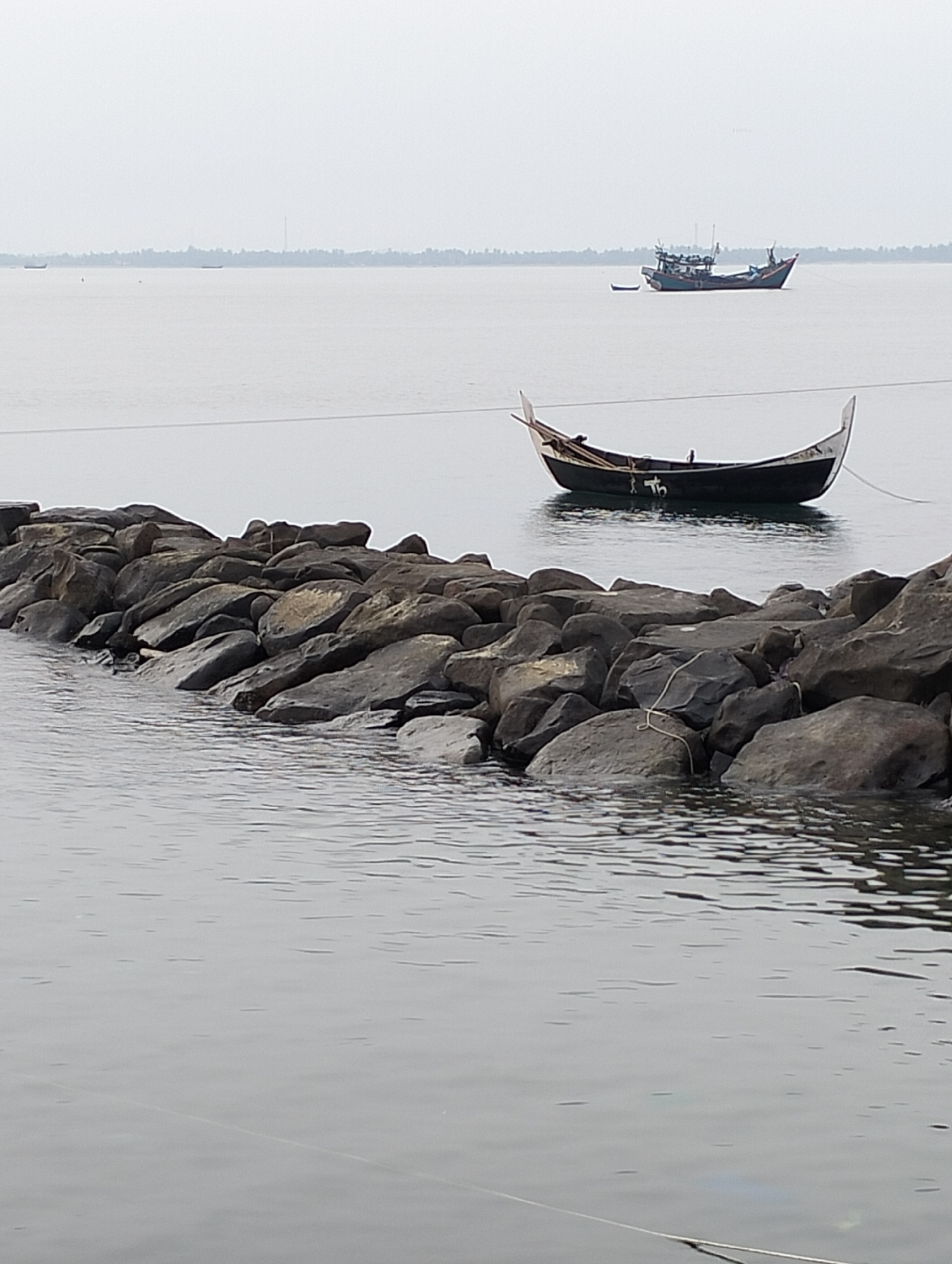 The image displays a coastal scene in Lhokseumawe, Indonesia. In the foreground, a man-made breakwater constructed from large, dark grey, irregularly shaped stones extends into the calm, grey water from the left. A black or dark-hulled wooden boat, featuring a distinctive high, pointed white bow and stern, is moored in the mid-ground, floating behind the rock barrier. A light-colored rope extends from this boat, crossing over the stones. Markings, possibly "th" or "fb," are visible on the boat's side. Further in the distance, a larger, multi-decked fishing vessel, primarily blue and dark-colored, is visible, accompanied by a smaller, light-colored skiff to its left. The horizon reveals a distant coastline with a visible treeline, consistent with palm trees, and two slender, tall structures, possibly industrial towers. The sky is uniformly light grey, indicating an overcast or hazy condition. No people are present in the scene.