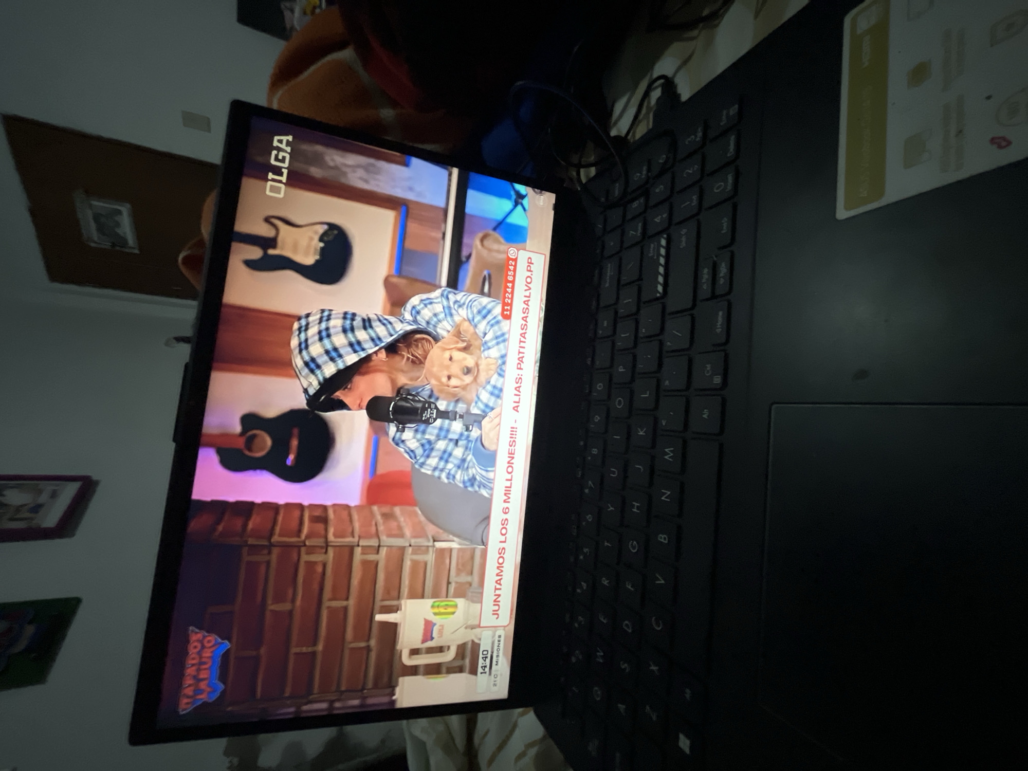 The image shows a laptop open on a bed. The laptop screen displays a broadcast from "OLGA" featuring a woman in a blue and white plaid hoodie holding a golden retriever puppy. A microphone is positioned in front of the woman. The backdrop behind them appears to be a brick wall and two electric guitars. A graphic on the screen reads "JUNTAMOS LOS 6 MILLONES!!!! - ALIAS: PATITASASALVO.PP". Below this, a time stamp of "14:40" and temperature reading of "21C" are visible. The laptop itself is black with a visible keyboard and trackpad. The background of the room is dimly lit and shows a door with a framed picture on it, and a portion of a bed with blankets. The presence of the "OLGA" broadcast suggests a possible location in Santa Ana, Venezuela, where this media outlet is known to operate.