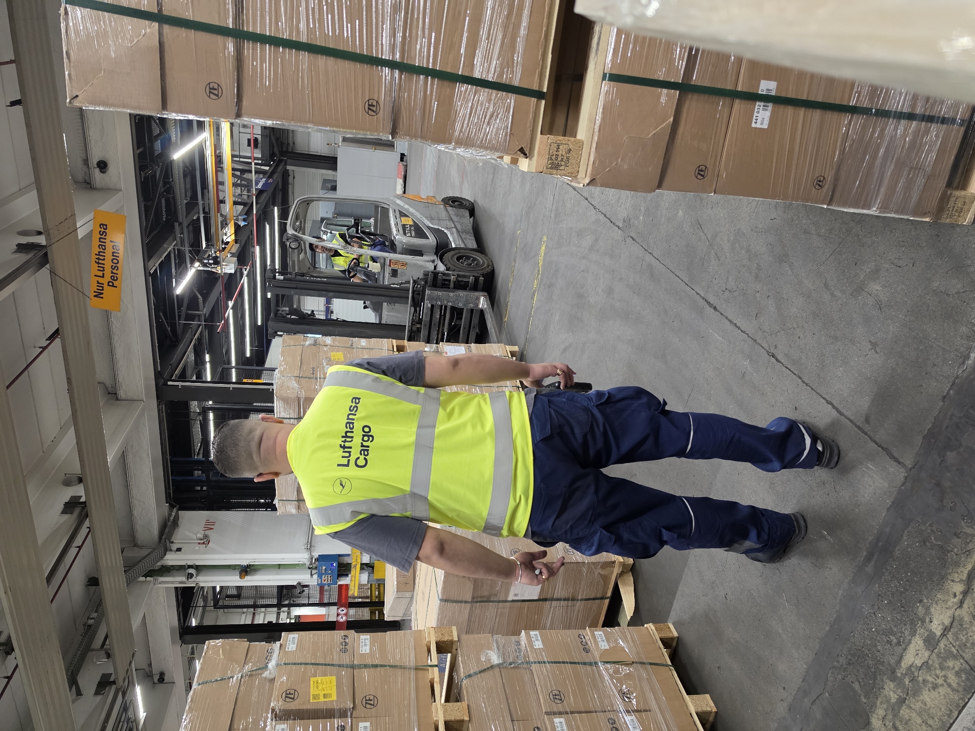 The image depicts a warehouse environment, likely within Frankfurt Airport (FRA) given the "Lufthansa Cargo" branding visible on a worker's vest. The scene is dominated by stacks of boxed goods, wrapped in plastic and secured with straps, positioned on wooden pallets. The flooring is a grey concrete surface with painted yellow lines.
In the foreground, a person wearing a high-visibility yellow vest with "Lufthansa Cargo" printed on it and dark blue work trousers is facing away from the camera, walking. The vest has reflective stripes. They are holding a small black object, possibly a scanner or radio, in their right hand.
In the mid-ground, a forklift is being operated by another individual also wearing a high-visibility vest. The forklift is a silver and grey model with the brand "STILL" visible. The forklift appears to be engaged in moving or positioning cargo.
Above the forklift, a yellow sign with black German text reads "Nur Lufthansa Personal" which translates to "Lufthansa Personnel Only".
The background shows further warehouse infrastructure, including elevated conveyor systems, lighting fixtures, and large industrial doors or loading bays. The overall atmosphere is that of an active logistics or shipping facility.