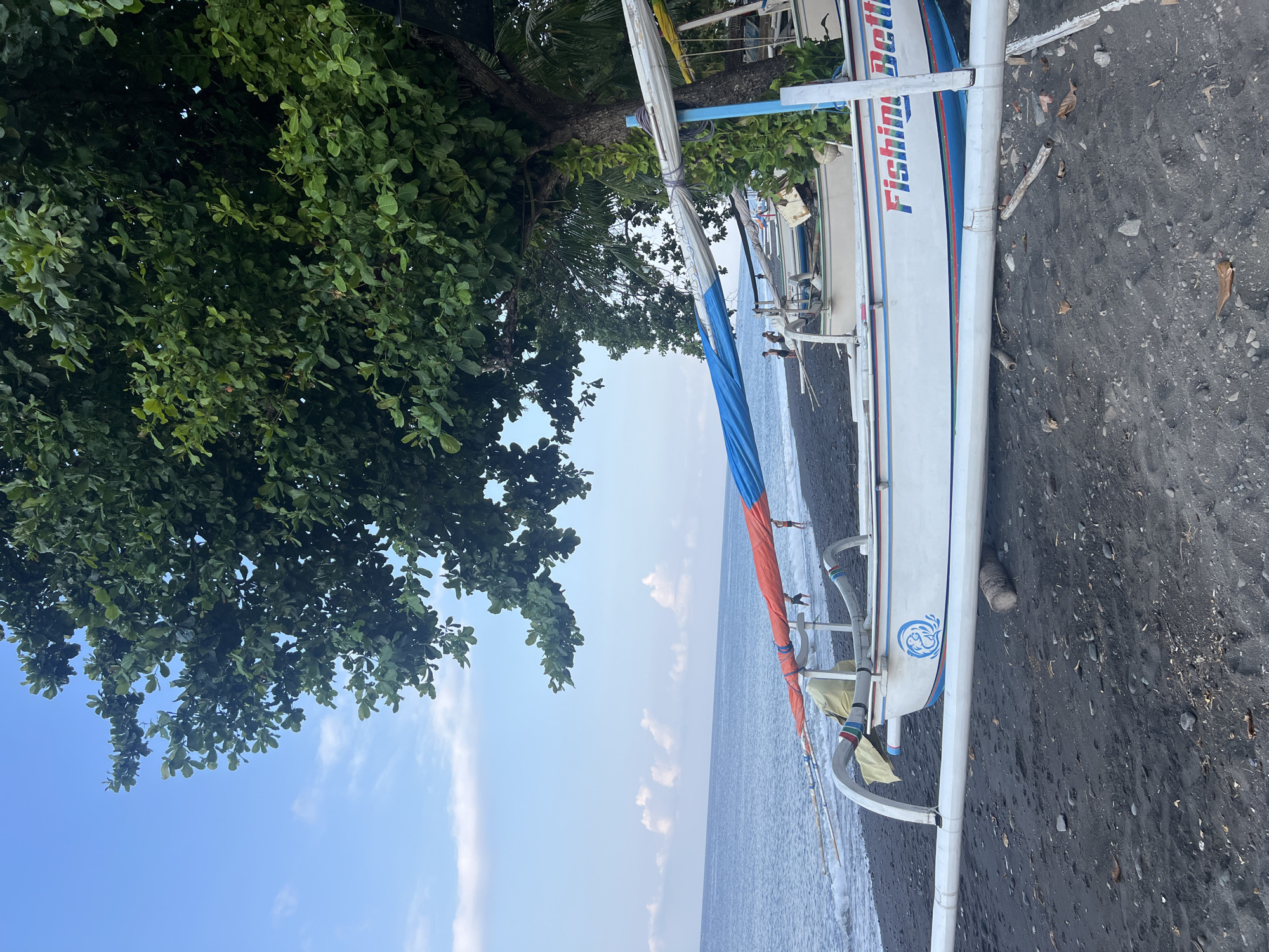 The image depicts a beach scene in Abang, Indonesia, under a bright blue sky with scattered white clouds. In the foreground, a traditional Indonesian outrigger boat, painted white with blue and red trim, rests on black volcanic sand. The boat is inscribed with the words "Fishing Bettie". The boat has outriggers extending from its sides, and a colorful sail, partially furled, is visible. A large tree with dense green foliage overhangs the beach from the right side of the frame. The ocean is calm, with gentle waves lapping at the shore. In the distance, a few people can be seen wading in the shallow water near the shoreline. The overall environment is serene and tropical.