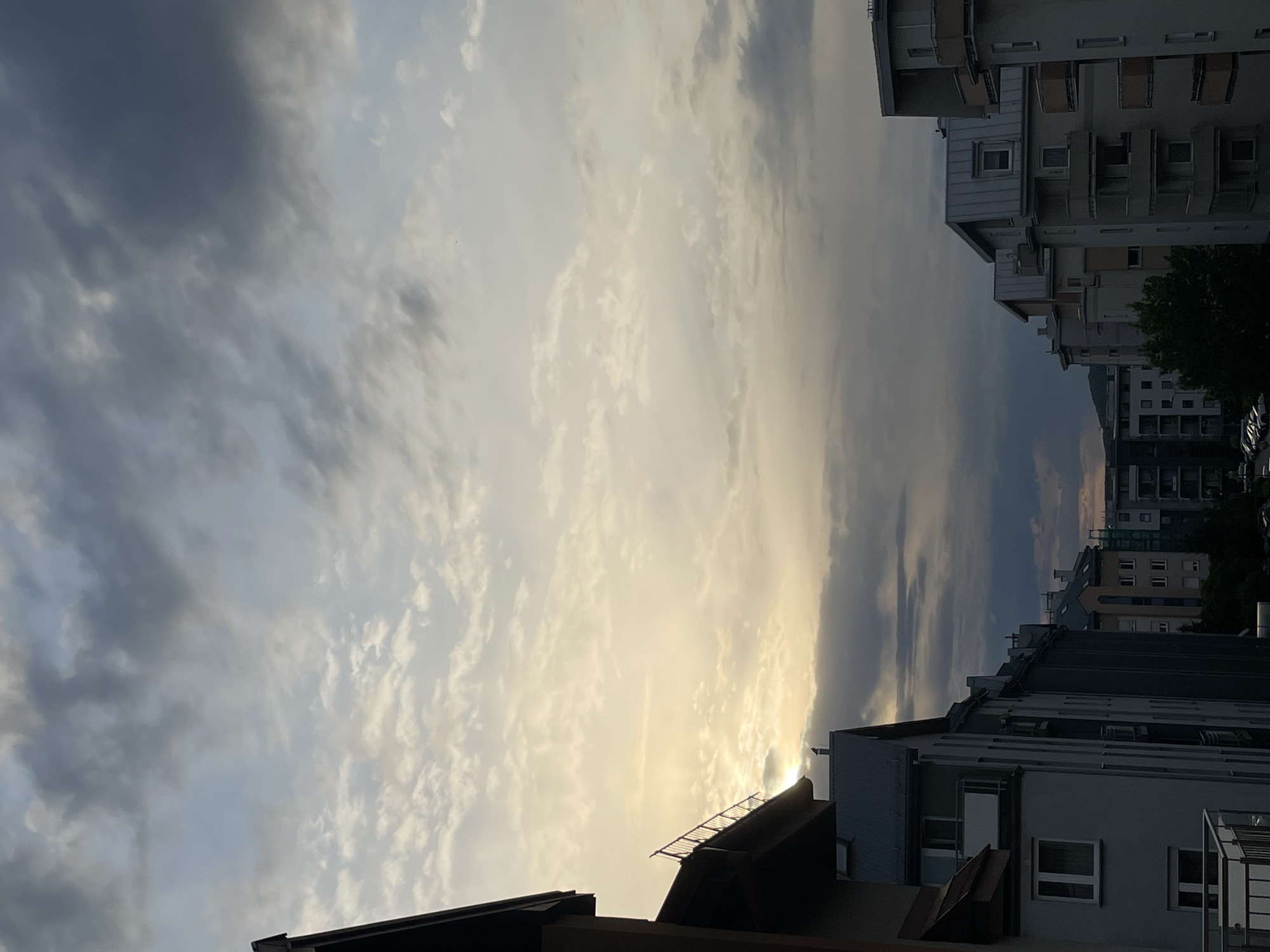 The image depicts a cityscape under a cloudy sky, possibly during sunrise or sunset. In the foreground, rooftops of buildings are visible, suggesting an urban setting. The sky is filled with a mix of dark and light clouds, with a bright area indicating the position of the sun. In the background, taller buildings are present, some with visible balconies and windows. The scene appears to be in a city, but the specific location is not discernible from the image alone.