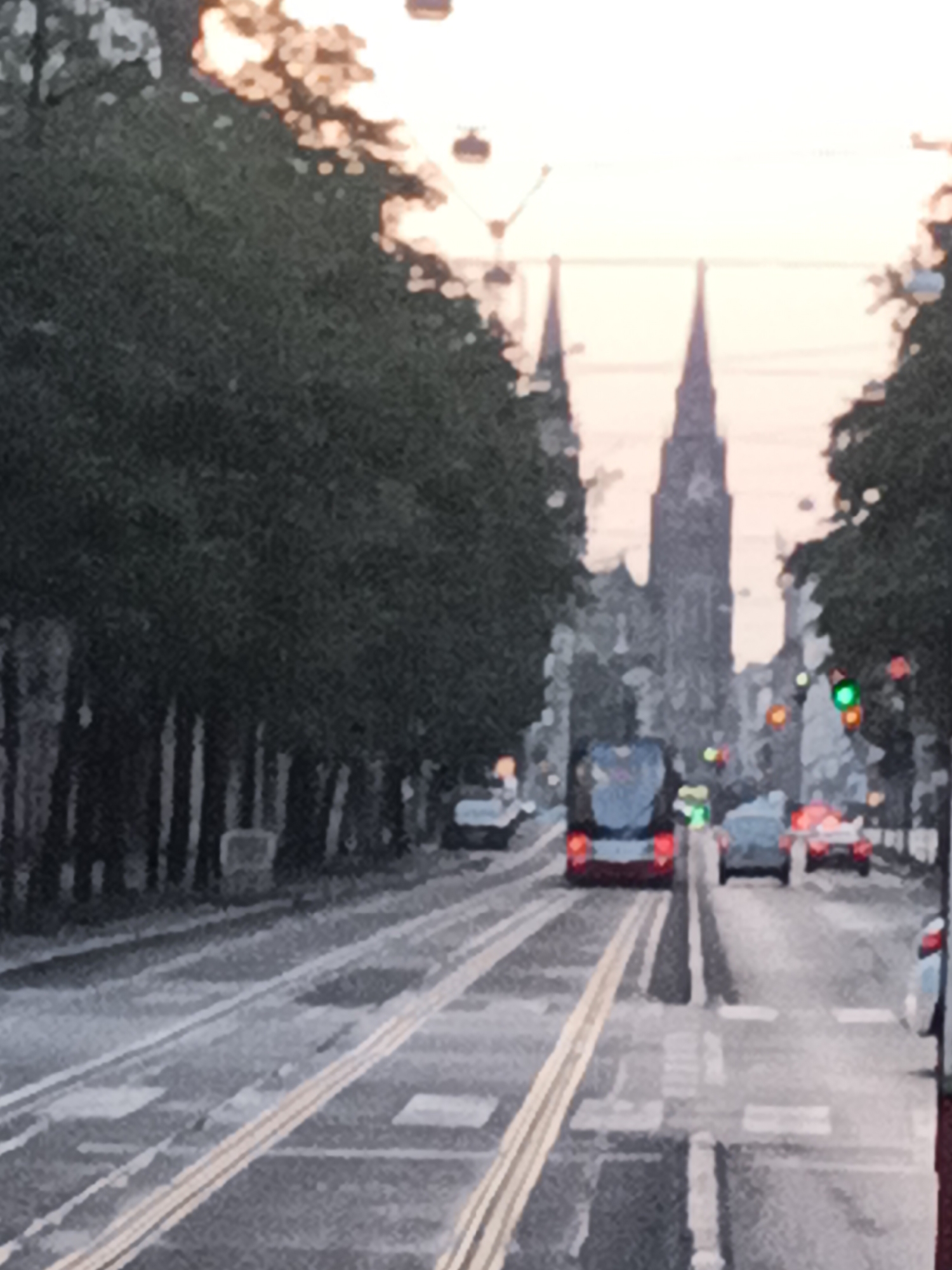 This image shows a street scene in Prague, Czechia, likely during the day with a hazy sky. The foreground features tram tracks running along the asphalt road. On the left, a line of trees with dark foliage creates a visual boundary. In the midground, a red and blue tram is seen moving away from the viewer. Several cars are also present on the road, including a white car on the right side and red vehicles further down. The background is dominated by the spires of two prominent buildings, characteristic of Prague's architecture, possibly churches or cathedrals. Traffic lights are visible, with a green light illuminated on one. The overall impression is one of a busy urban environment with historical structures.