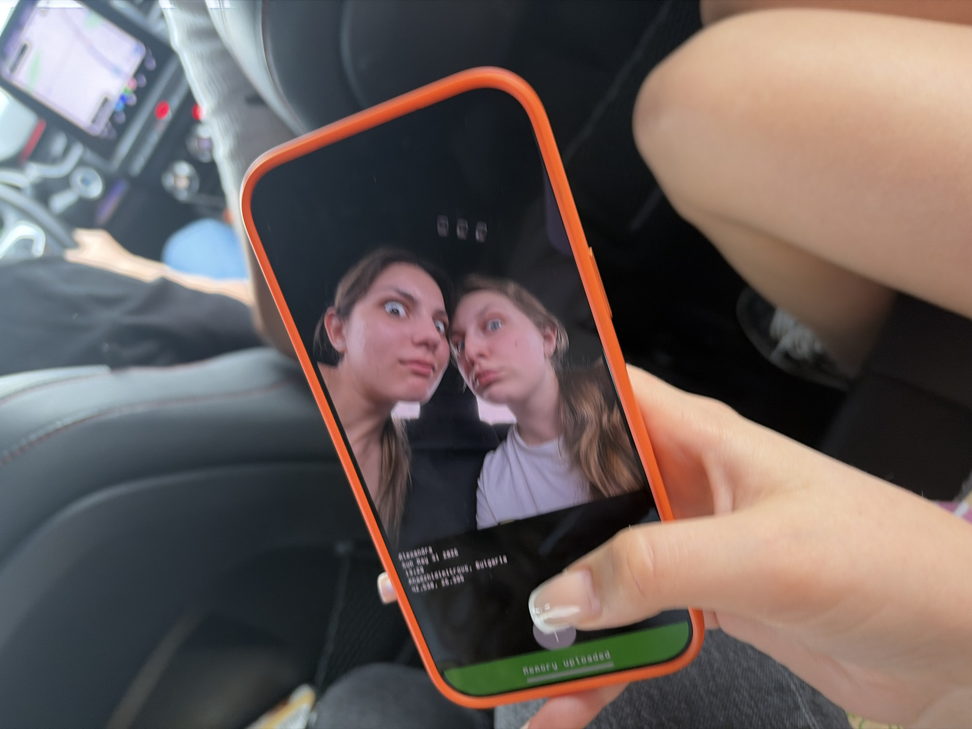 A close-up image displays a person holding an orange-cased smartphone, which displays a photograph of two young women making silly faces. The metadata on the phone screen indicates "aleksandra," "Sun May 31 2020," "15:25," "Aleksandarovo, Bulgaria," and coordinates "42.9808, 25.3890." The background shows the interior of a car, specifically the dashboard and passenger seating area. The person holding the phone has their thumb near the bottom of the screen, seemingly about to press a button that says "Memory Uploaded." The environment suggests they are sitting in a car, possibly within the city of Chokoba, Bulgaria.