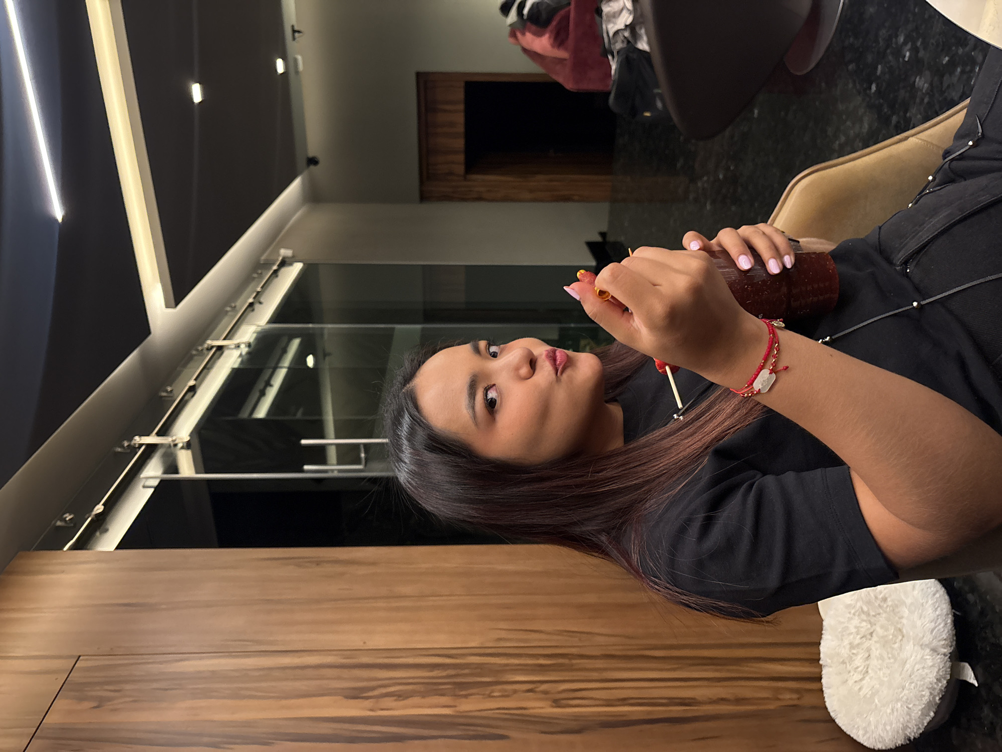 A medium shot of a woman sitting indoors, turned to the side, holding a bottle and skewers of food in her hands. The setting appears to be a modern building, possibly a restaurant or a home with large windows and contemporary lighting. The woman has dark hair and is wearing a black shirt. She is wearing a red bracelet on her left wrist. The floor is a dark color, and furniture is visible in the background. Based on the modern architectural style and the person's appearance, the image may have been taken in San Bernardino Tlaxcalancingo, Mexico, but there are no definitive clues in the image to confirm that location.