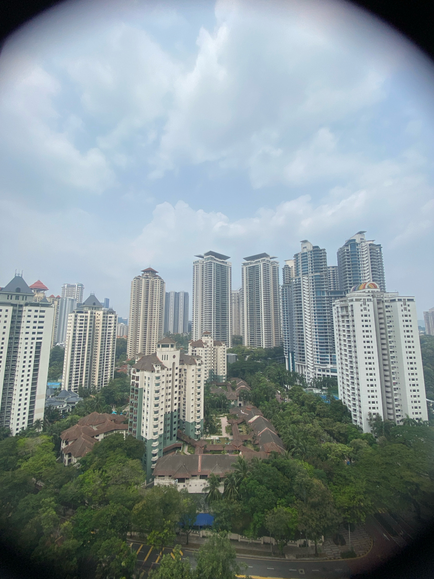 The image shows a high-angle view of a dense urban landscape in Kuala Lumpur, Malaysia, characterized by a mix of modern high-rise apartment buildings and lower-rise residential structures interspersed with lush greenery. The foreground and edges of the image are darkened by a vignetting effect, suggesting it was taken through a lens or aperture.
Dominating the scene are numerous residential skyscrapers, varying in architectural style and height, with clean lines and numerous windows. Many of these buildings are clad in white or light-colored materials, reflecting the overcast sky. Some feature prominent balconies or terraced elements. Among the taller buildings, there are also clusters of mid-rise apartment complexes, appearing to have a more traditional design with sloping tiled roofs and white or cream facades.
A significant amount of mature tropical foliage, including tall trees and dense undergrowth, is integrated throughout the residential areas, creating a contrast between the built environment and nature. This green space is particularly evident surrounding the lower-rise buildings, suggesting a planned community that prioritizes green living.
The sky is overcast with diffused light, indicating a cloudy or hazy day. The overall impression is one of a densely populated, well-developed residential district within Kuala Lumpur, where high-rise living is complemented by substantial green spaces. There are no visible people or specific activities depicted in the image, focusing solely on the urban and natural landscape. The presence of roads and pathways suggests underlying infrastructure for the residential areas.