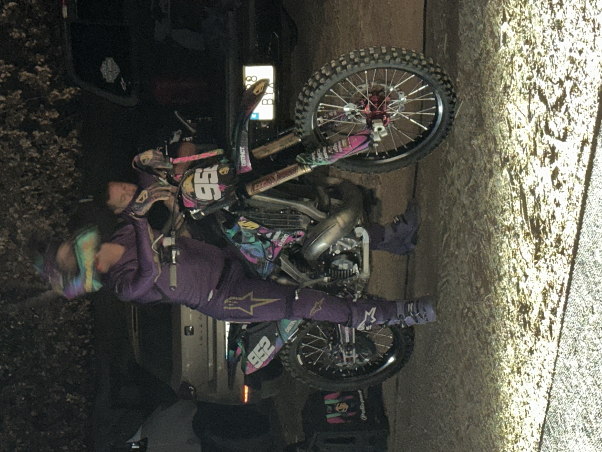 A person is posing on a dirt bike in the city of San José Del Valle, Spain. The person is wearing a purple motocross suit and helmet, with colorful graphics on the bike. The background shows a dark outdoor area with trees and vehicles. The ground is covered in gravel and illuminated by a bright light source.