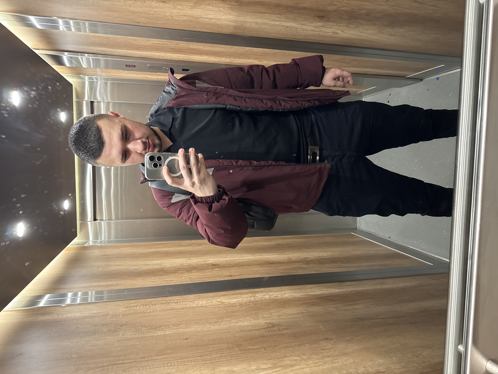 A man is taking a selfie in an elevator. He is wearing a dark-colored shirt, black pants, and a maroon and gray jacket. He is holding a silver-colored smartphone. The elevator has wooden-paneled walls and a silver door. The location is not discernible from the image. There is no indication of the city being Sofia, Bulgaria.