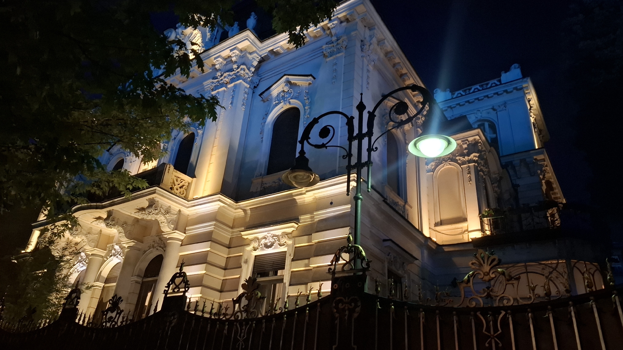 This image depicts the Ivan Vazov National Theatre in Sofia, Bulgaria, at night. The theatre is illuminated by warm yellow and cool blue lights, highlighting its ornate facade and architectural details. A wrought-iron fence runs along the foreground, partially obscuring the view of the building. A vintage-style lamppost with a glowing green bulb stands in front of the theatre, casting a soft light on its surroundings. Trees with dark green leaves are visible on the left side of the image, framing the theatre and adding a touch of nature to the urban scene. The night sky is dark, with a few faint clouds visible in the upper right corner. The overall atmosphere is one of grandeur and timeless beauty, with the interplay of light and shadow creating a dramatic and evocative mood.