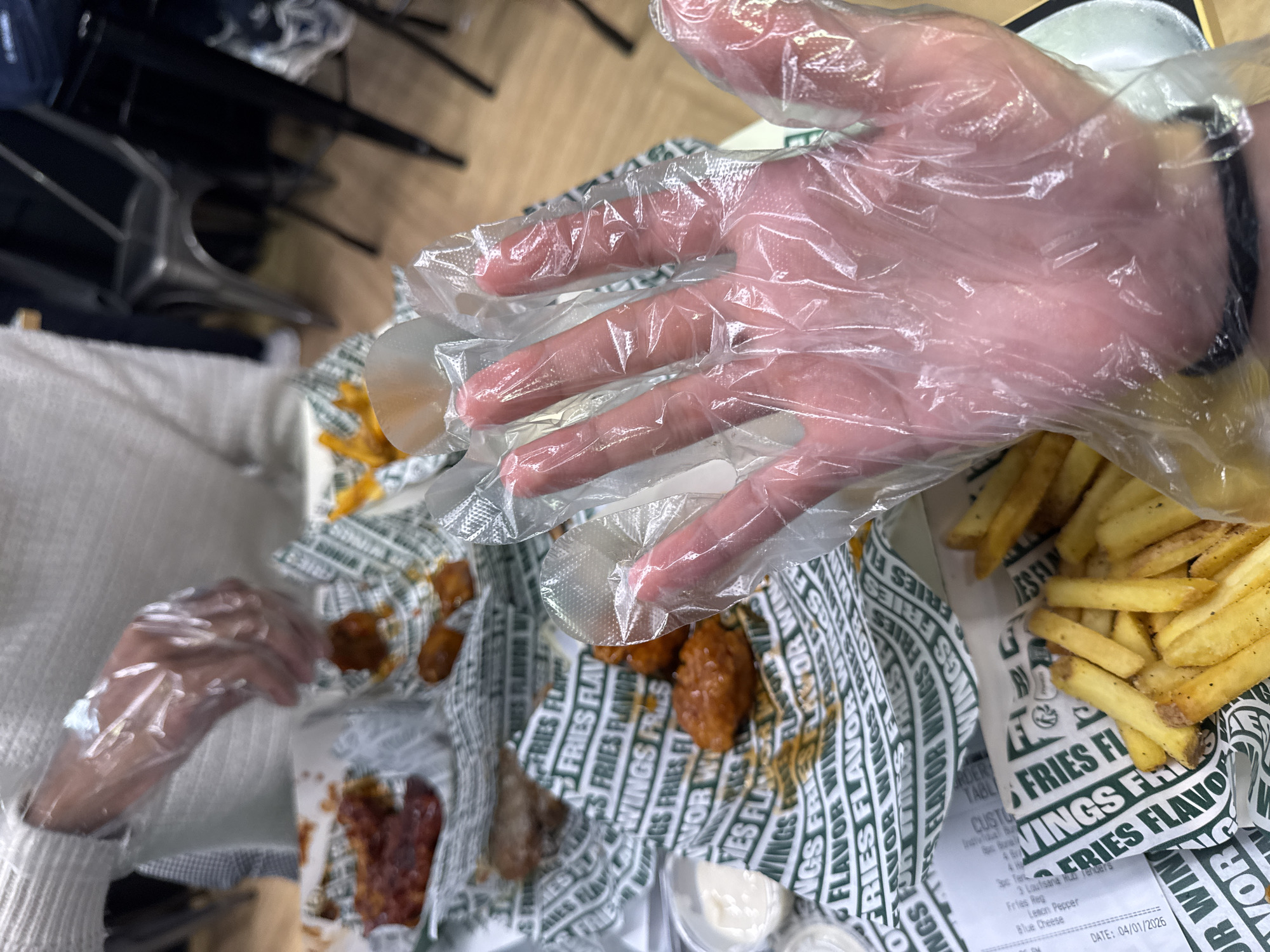 The image depicts a person eating at what appears to be a Wingstop restaurant, recognizable by the branded paper lining the trays holding their meal. The meal consists of chicken wings with various sauces and french fries. The person is wearing clear plastic gloves on both hands, presumably to eat the wings without getting their hands messy. A receipt is visible with the order details and the date, April 1st, 2026. The location is unknown, but given the receipt details, it is likely not in Paris, France. The scene takes place at a table, with chairs visible in the background.