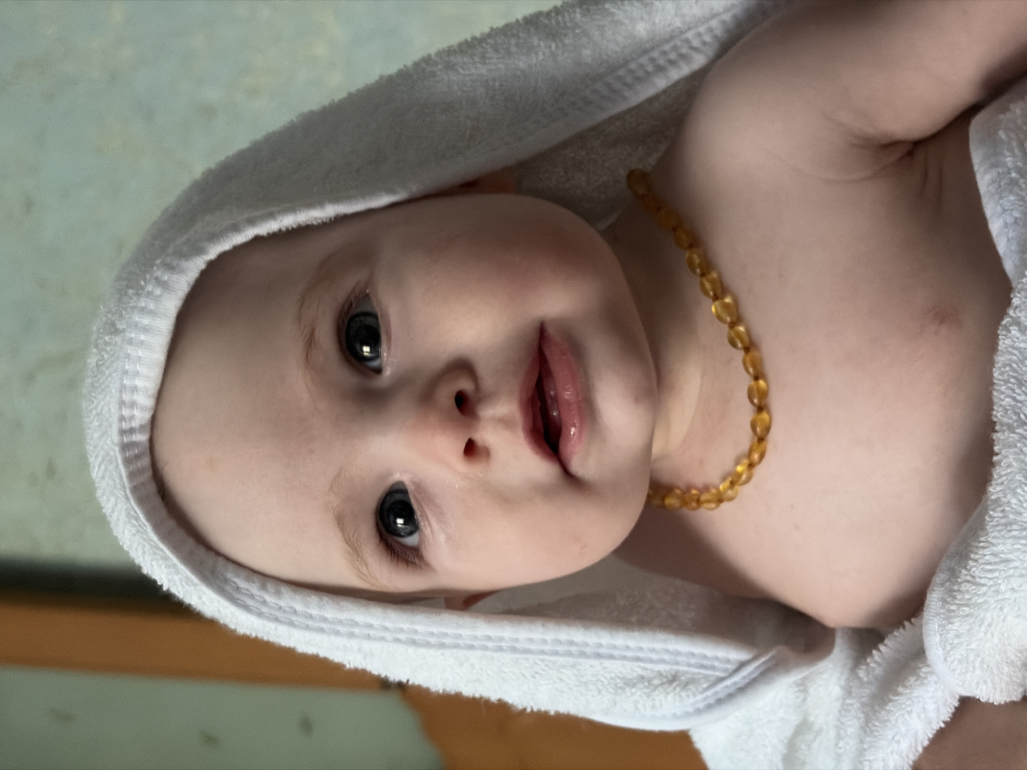 The image shows a close-up, sideways portrait of a baby wrapped in a white towel. The baby has fair skin, dark eyelashes, and appears to be looking directly at the camera with a curious expression. The baby is wearing a necklace of amber beads. The background is a soft, blurred green and beige, suggesting an indoor setting, possibly a bathroom or nursery. The lighting appears natural and soft, with no harsh shadows, implying it might be daytime. The overall mood is intimate and peaceful. There is no visible text in the image.