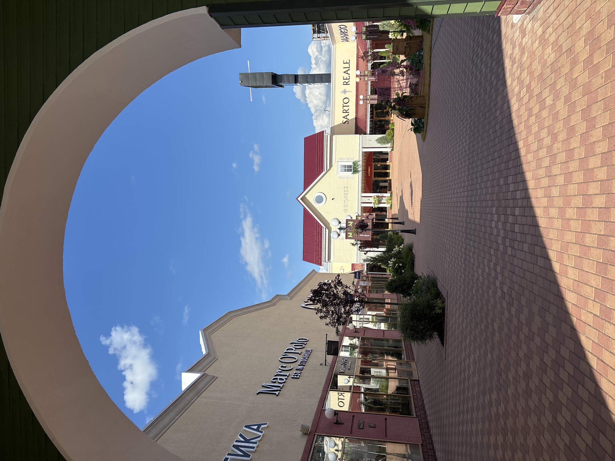 The image depicts an outdoor shopping area with a paved walkway and buildings on either side. The walkway is constructed of red and beige interlocking pavers, with a prominent shadow cast across it from the left. On the left side of the walkway, there is a building with a light tan facade, featuring large windows and illuminated signs for "Marc O'Polo" and "ОТЯ". The windows of the shops are reflective, showing glimpses of merchandise inside. A dark-leaved tree stands in front of the Marc O'Polo store.
On the right side of the walkway, there are buildings with yellow facades and red tiled roofs. One building has a sign that reads "12 STOREEZ" above its entrance. Further down, another building displays signs for "SARTO REALE" and "MANGO". In front of these buildings, there are decorative displays with plants and what appear to be vintage suitcases.
The sky above is bright blue with scattered white clouds. In the distance, a tall, dark tower-like structure is visible against the sky. The scene appears to be a commercial district or outlet mall.