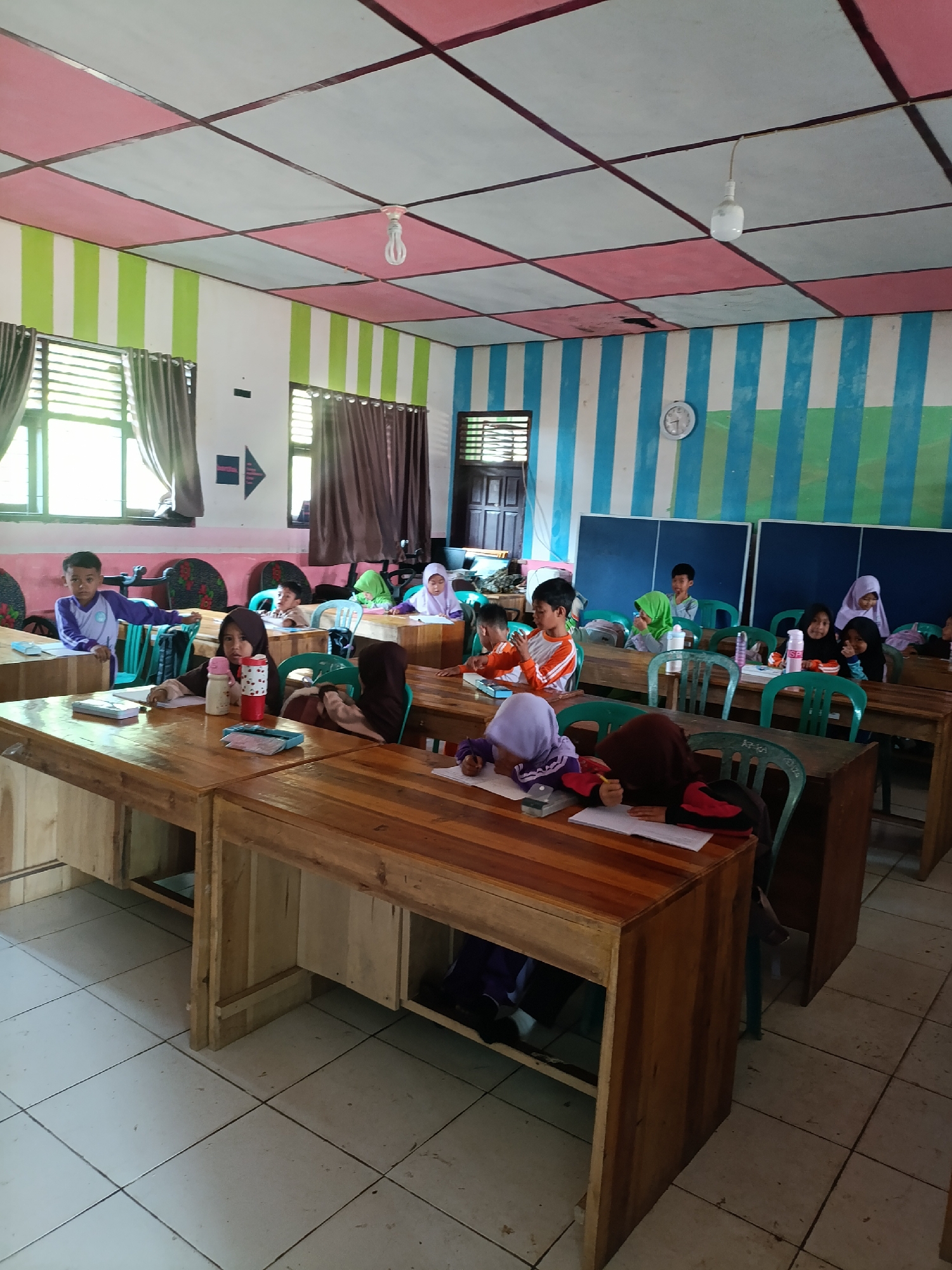This is a medium-shot, vertical-frame photograph taken inside a classroom in the city of Sumberejo, Indonesia. The classroom features desks made of wood, green chairs, and tiled flooring. The walls are painted with alternating vertical stripes of blue and white, and the ceiling is a grid of pink and light blue squares. A simple light bulb hangs from the ceiling. Several children are seated at the desks, engaged in learning activities. Some are looking towards the front of the classroom, while others are focused on their books or papers. The overall atmosphere appears to be one of a typical school day.