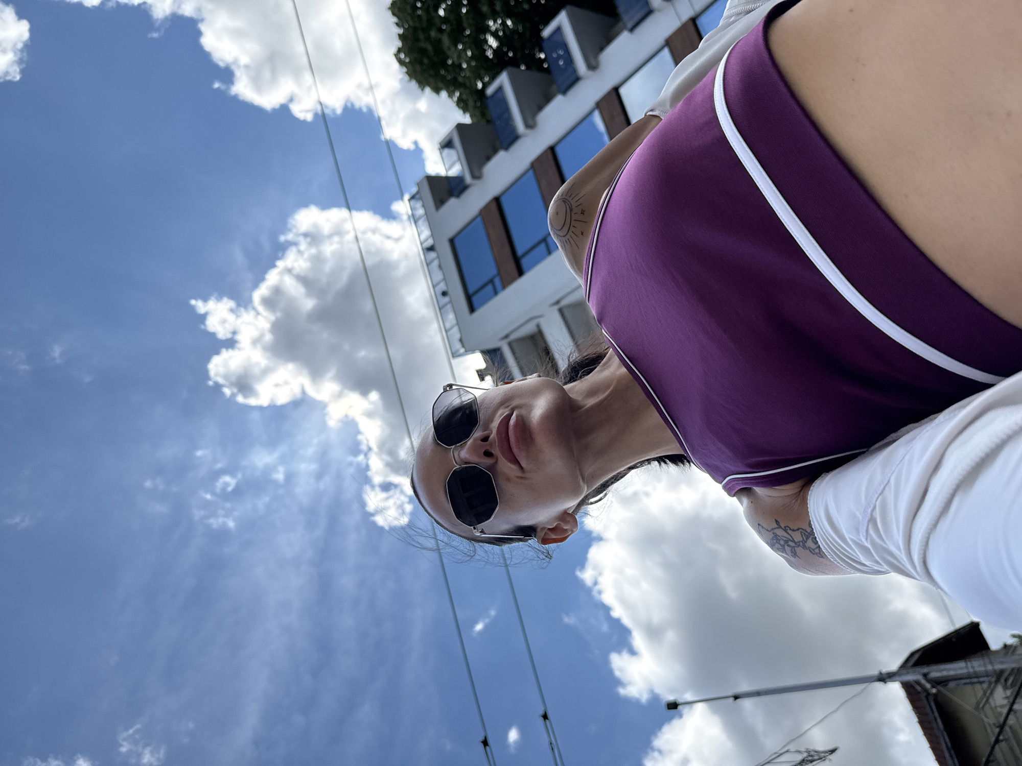 A low-angle, outdoor, waist-up shot features a woman wearing sunglasses and a purple tank top with white trim. She is looking up and to her left. Behind her, a modern building with blue-tinted windows and balconies is visible, along with trees and a blue sky with scattered white clouds. The scene is set in the city of Pleven, Bulgaria.