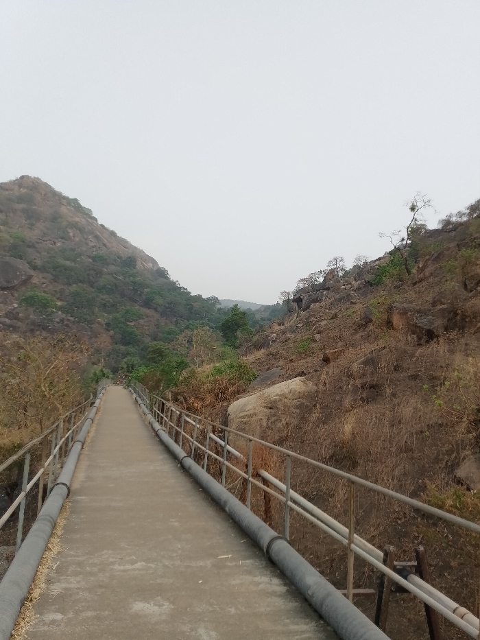 A narrow concrete pathway stretches forward, flanked by metal railings and large black pipes. On either side, hills rise, covered with dry vegetation and scattered trees. A massive boulder sits beside the path on the right. The sky is overcast, casting a muted light over the landscape. The scene evokes a sense of isolation and industrial functionality amidst natural terrain.