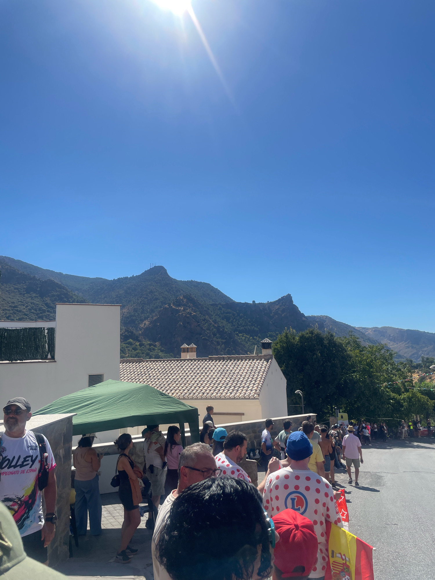 In Güéjar Sierra, Spain, under a clear blue sky with bright sunlight, a crowd gathers along a paved road leading uphill towards mountainous terrain. The mountains are covered in dense green vegetation, with some rocky outcrops and a structure with antennae visible on a distant peak. In the foreground, several people are lined up, some under a green tent. One man in the foreground wears a white t-shirt with colorful graphics and the word "VOLLEY." Another person wears a white shirt with red polka dots and a blue "L" logo. A Spanish flag, red and yellow, is held by someone in the crowd. Buildings with tiled roofs and white walls are visible, along with a dark green fence or screen on one building. The overall scene suggests an outdoor event or gathering.