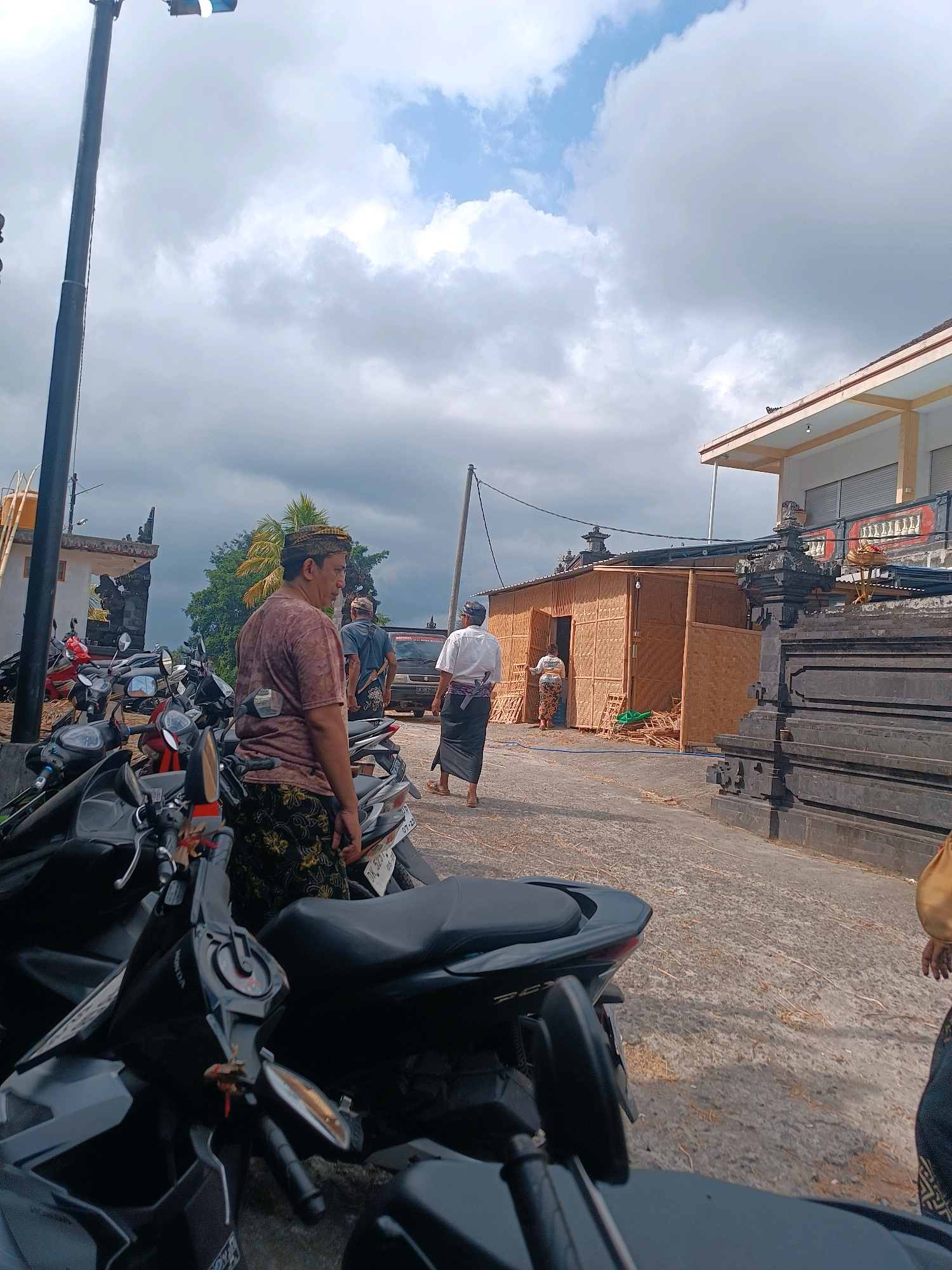 The image captures an outdoor scene in Selemadeg, Indonesia, under a cloudy sky. In the foreground, several parked motorcycles are visible, with one black scooter dominating the lower portion of the frame. To the left, a tall pole with a light fixture stands prominently. Several men, some wearing traditional Balinese attire including sarongs and head coverings, are present. One man, wearing a patterned sarong and a dark t-shirt, is in the middle ground, looking to the right. Behind him, a temporary structure made of woven bamboo is set up, with two people visible near its entrance. Further back, a modern building with a light-colored facade and dark shutters is visible. A stone wall or barrier with decorative elements is also present on the right side of the image. The ground is paved with concrete and scattered with dry organic material, possibly straw or leaves. The overall atmosphere suggests a cultural event or gathering.
