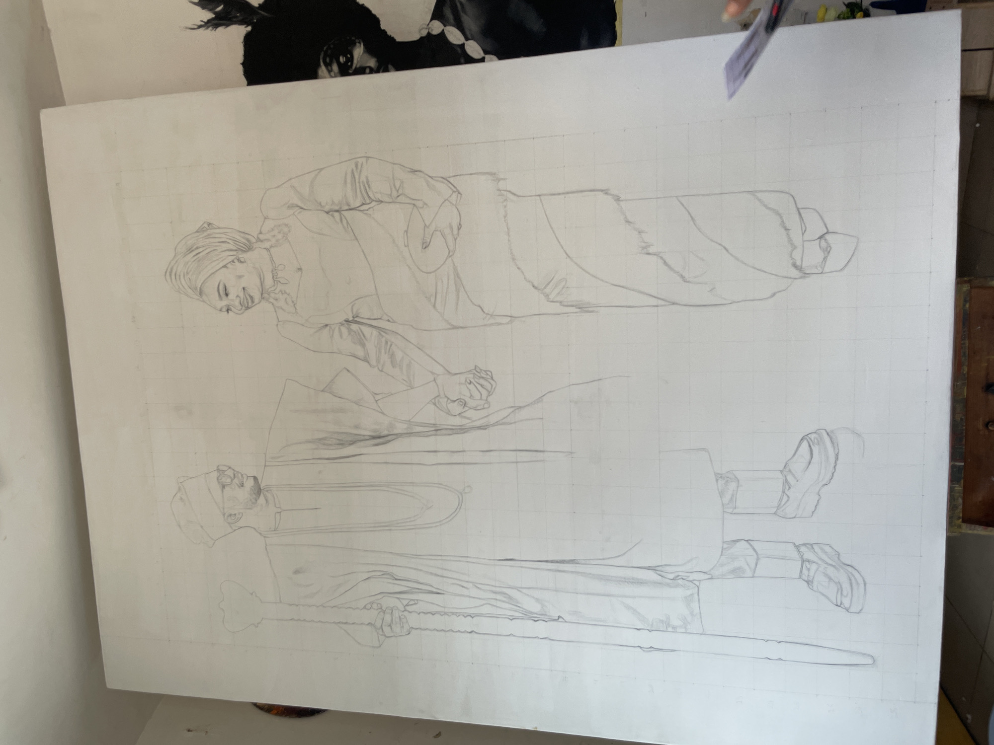 The image shows a large white canvas with a pencil sketch of two figures, likely people. The canvas is gridded with faint lines, suggesting a method for accurate scaling and transfer of the design.
The upper figure is reclining or lying down, possibly wearing a head covering (turban or headscarf) and what appears to be a fur-trimmed garment. Their hands are clasped with the figure below.
The lower figure is standing and appears to be holding a long, straight object, possibly a staff or weapon. They are wearing a long robe-like garment and shoes or boots.
The environment is an indoor space, likely a studio or workshop. There is a partial view of what appears to be another drawing or artwork in the upper left corner of the image. A ruler is visible in the upper right corner. The location is Unknown, Unknown.